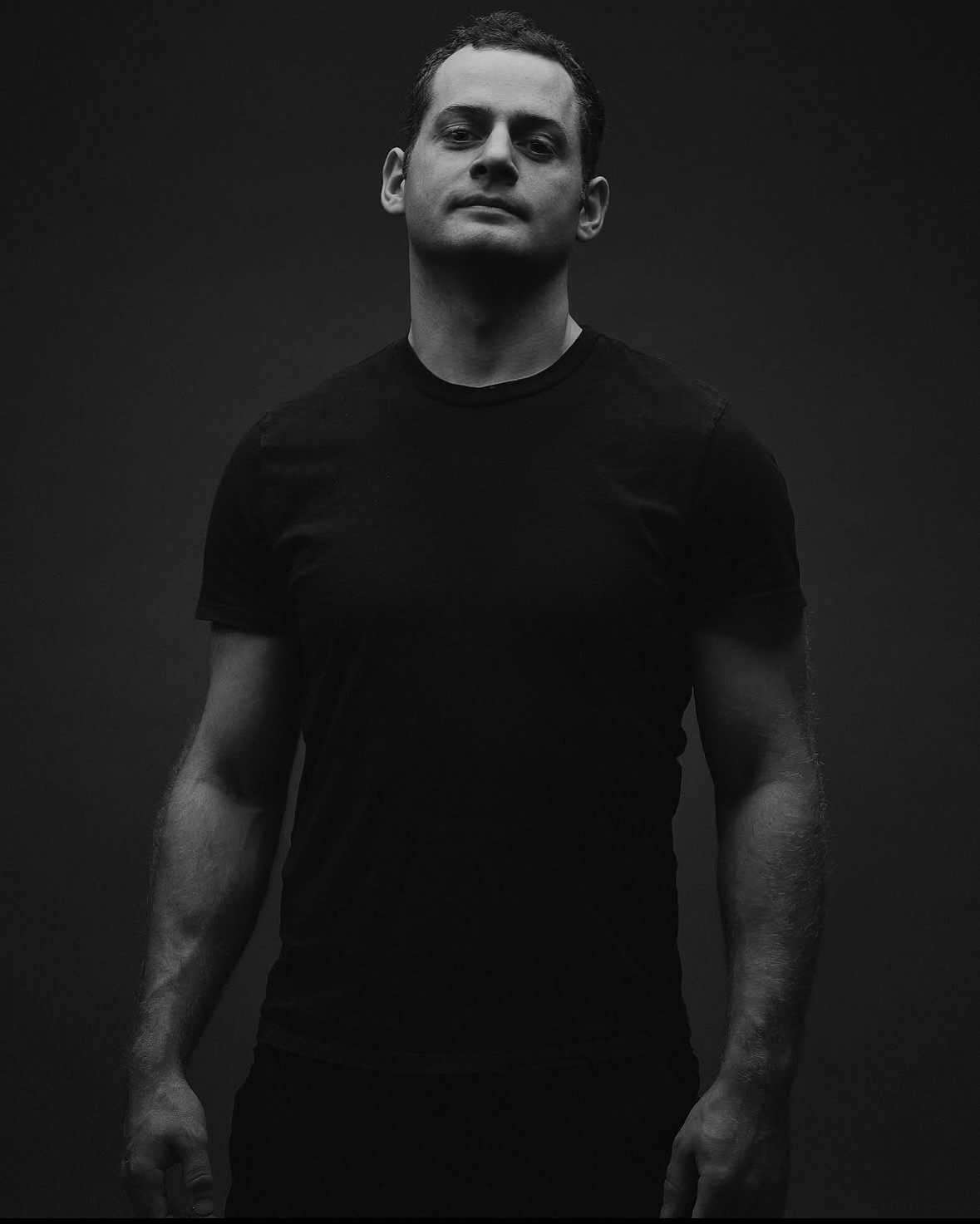 Man standing against dark background, wearing a black t-shirt, looking at camera with neutral expression.