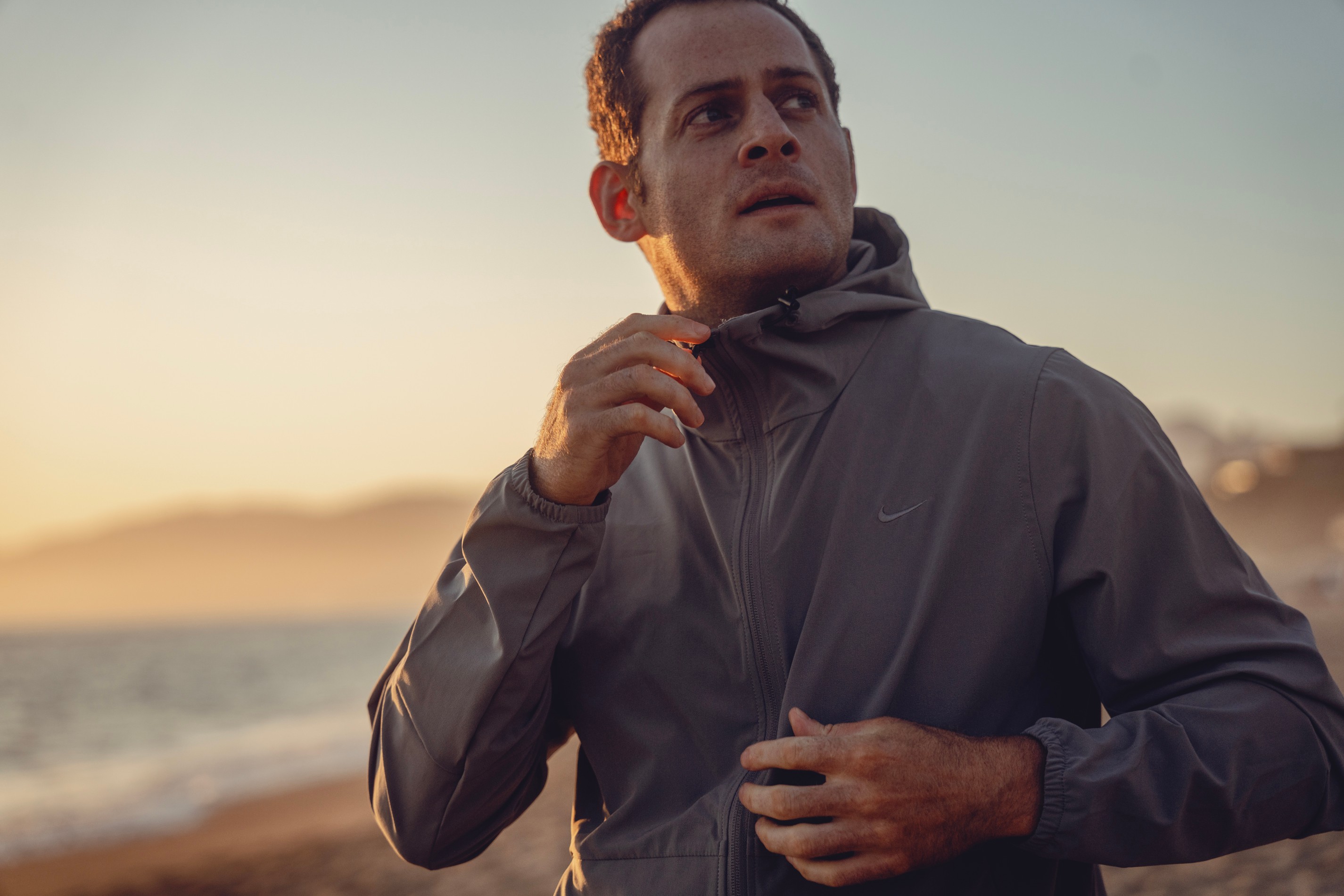 Man in gray jacket outdoors near water during sunset, looking to the side with hand near collar.