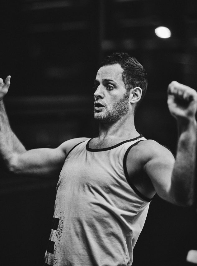 Man flexing muscles with arms raised, wearing a sleeveless shirt, in a gym setting.