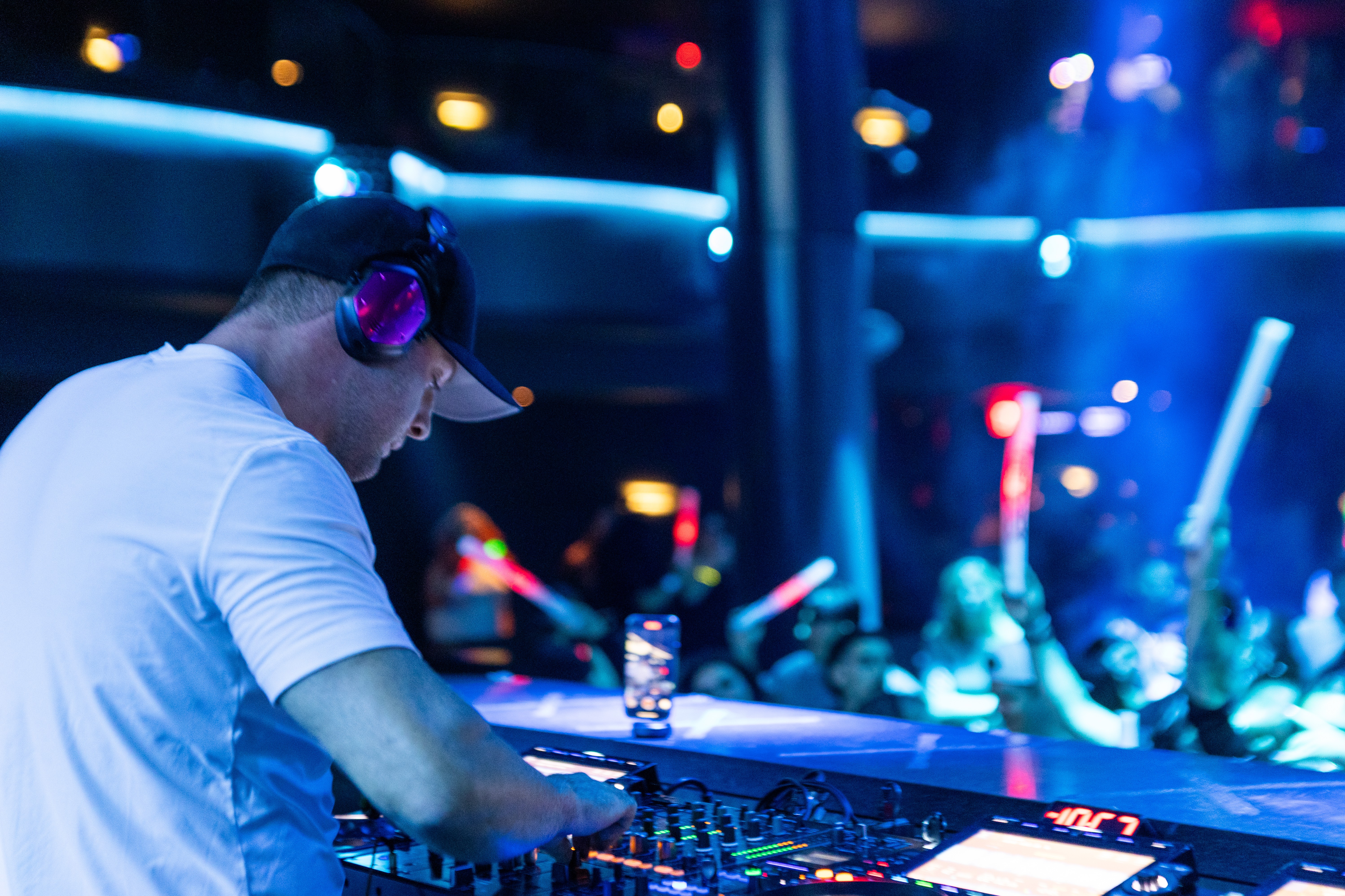 DJ wearing headphones and cap operates equipment in a nightclub with colorful lights and a crowd.