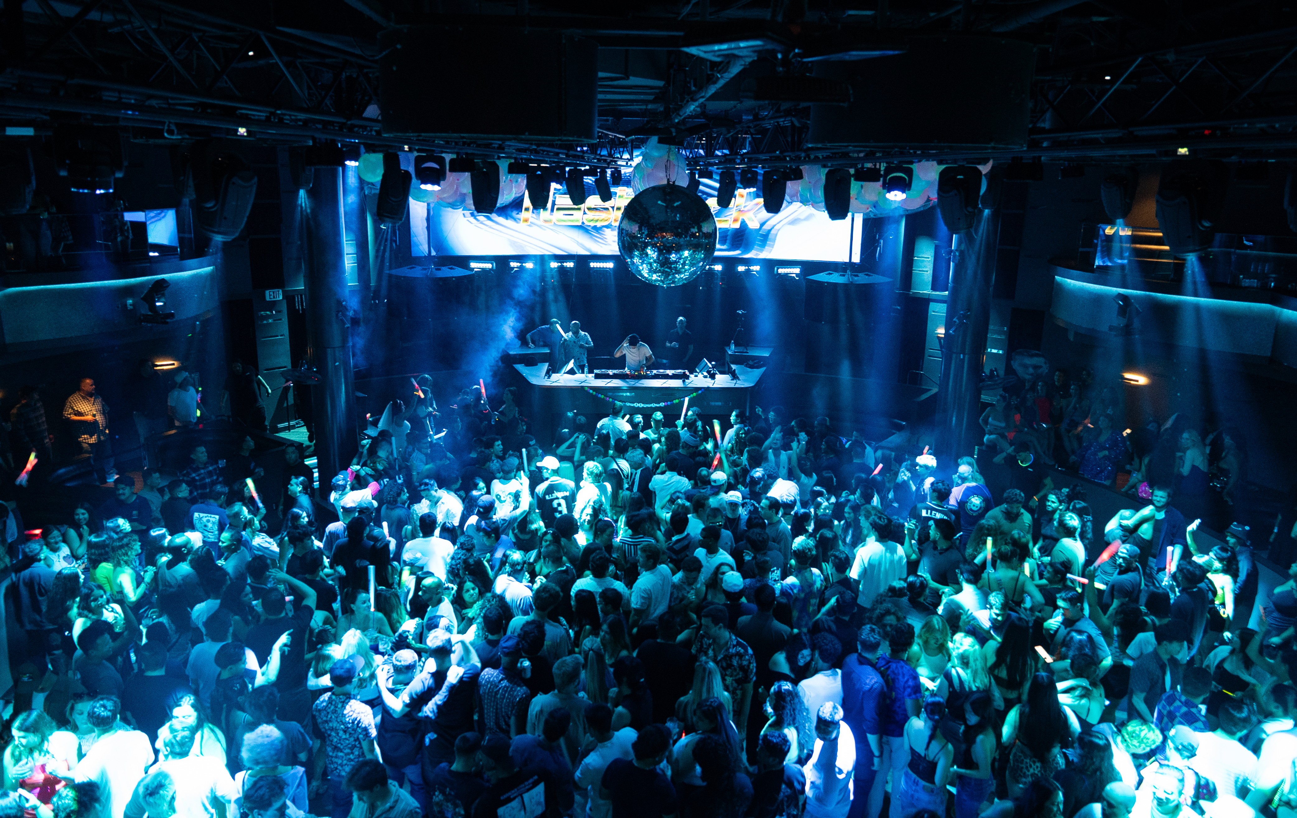 Crowd of people dancing in a nightclub with blue lighting and a DJ booth on stage.