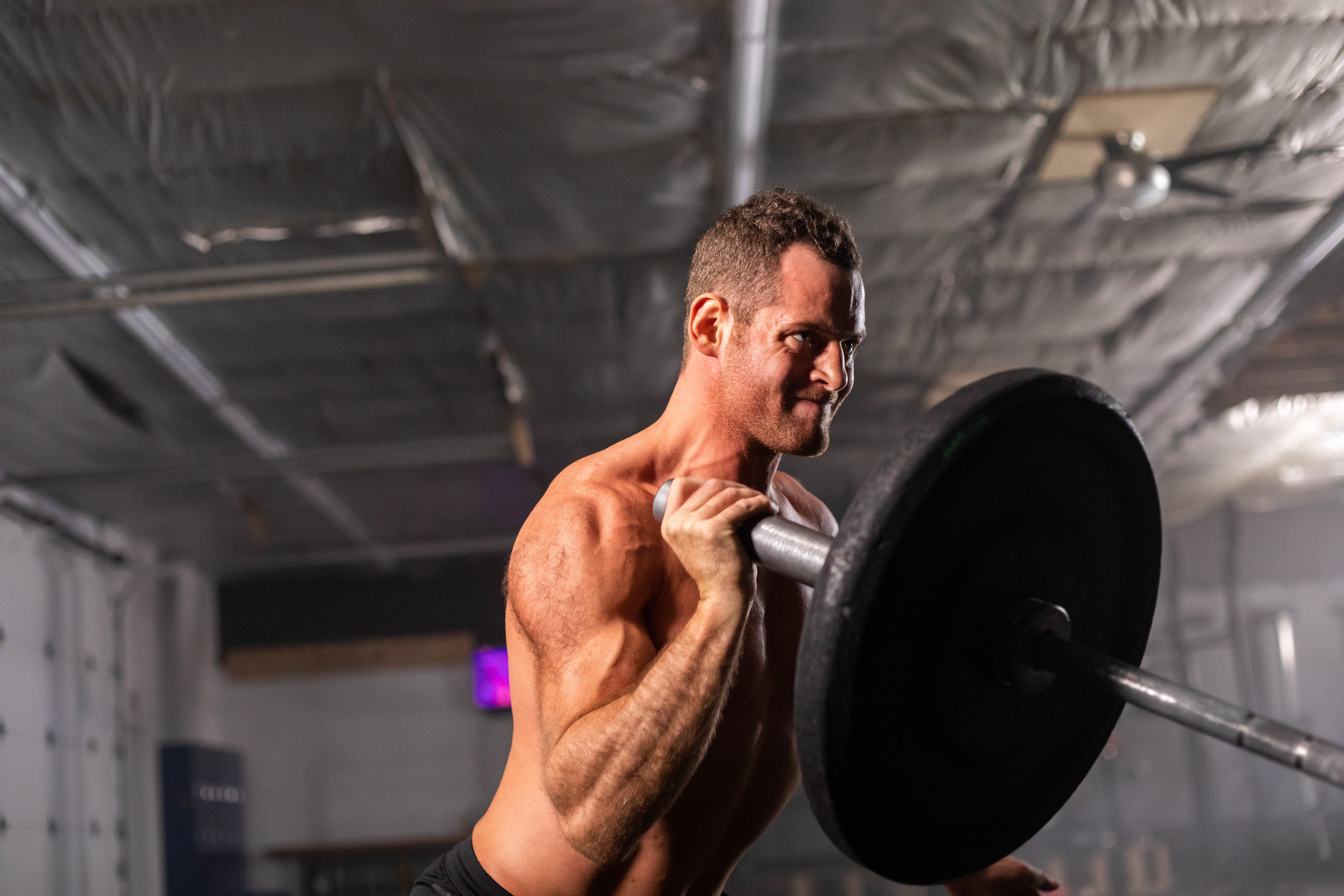 Man lifting a barbell with weights in a gym, shirtless, focused expression, ceiling with exposed pipes and lighting fixtures.