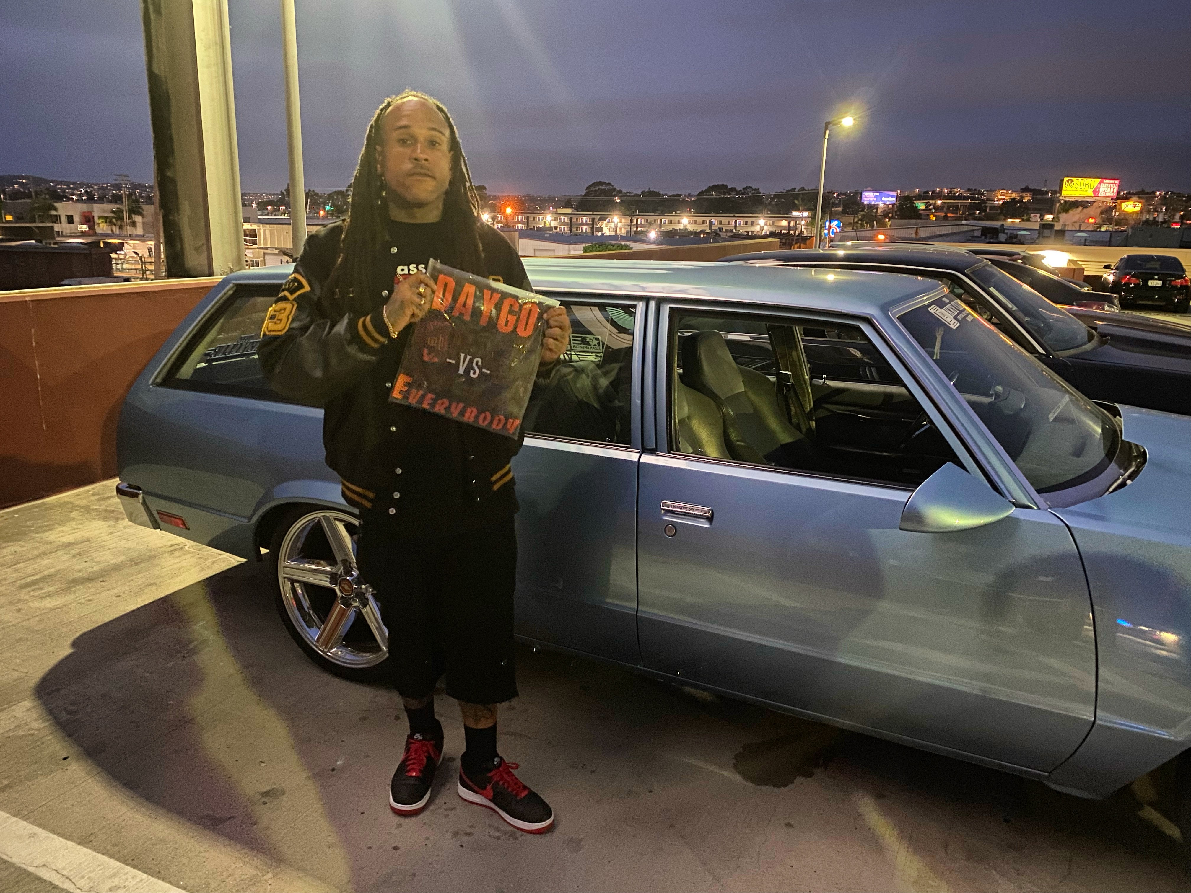Person with long hair holding a sign in front of a parked car on a city street at dusk.