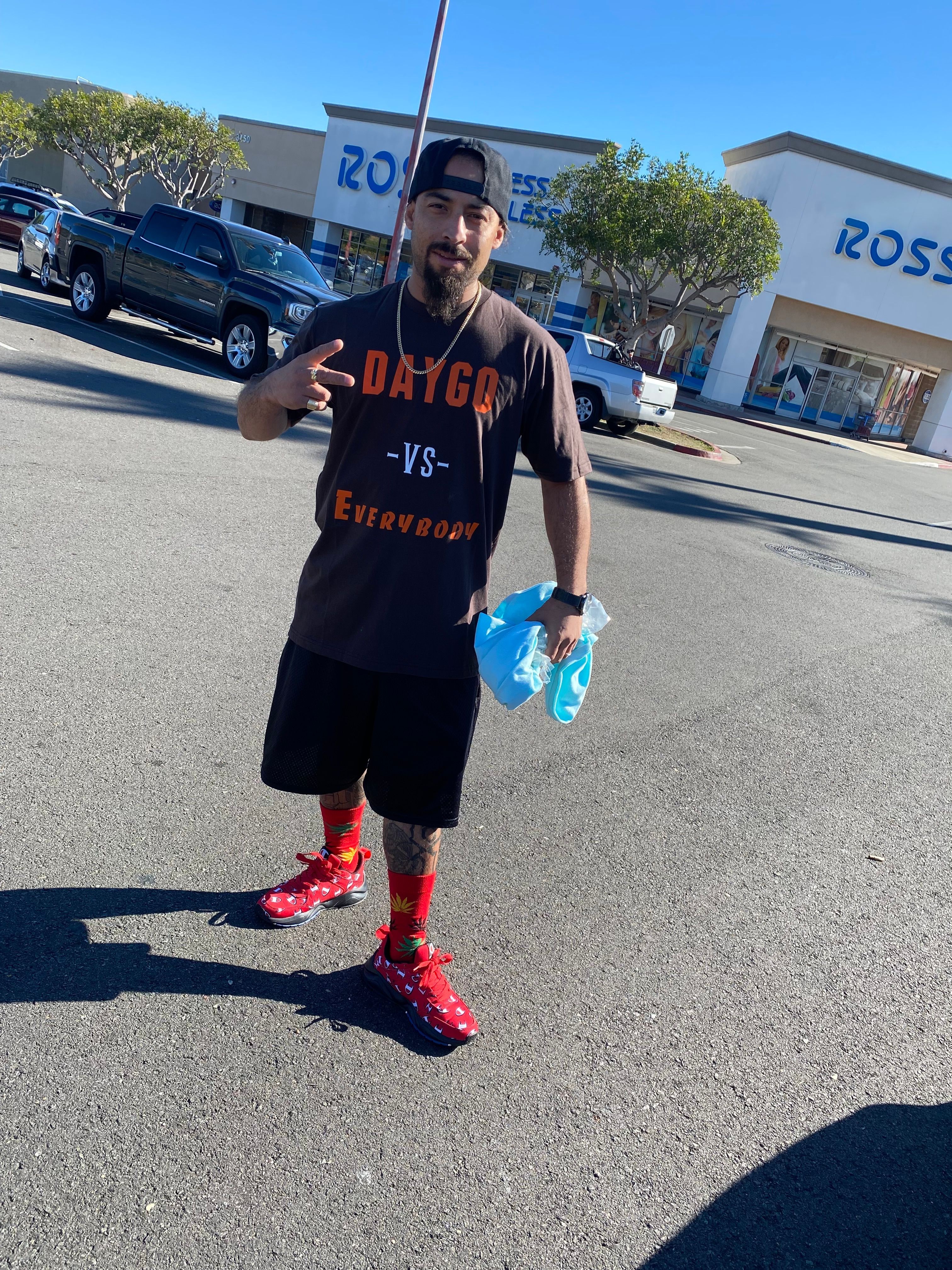 Young man standing on street holding blue water bottles, wearing black shorts, red shoes, and a black sleeveless shirt.