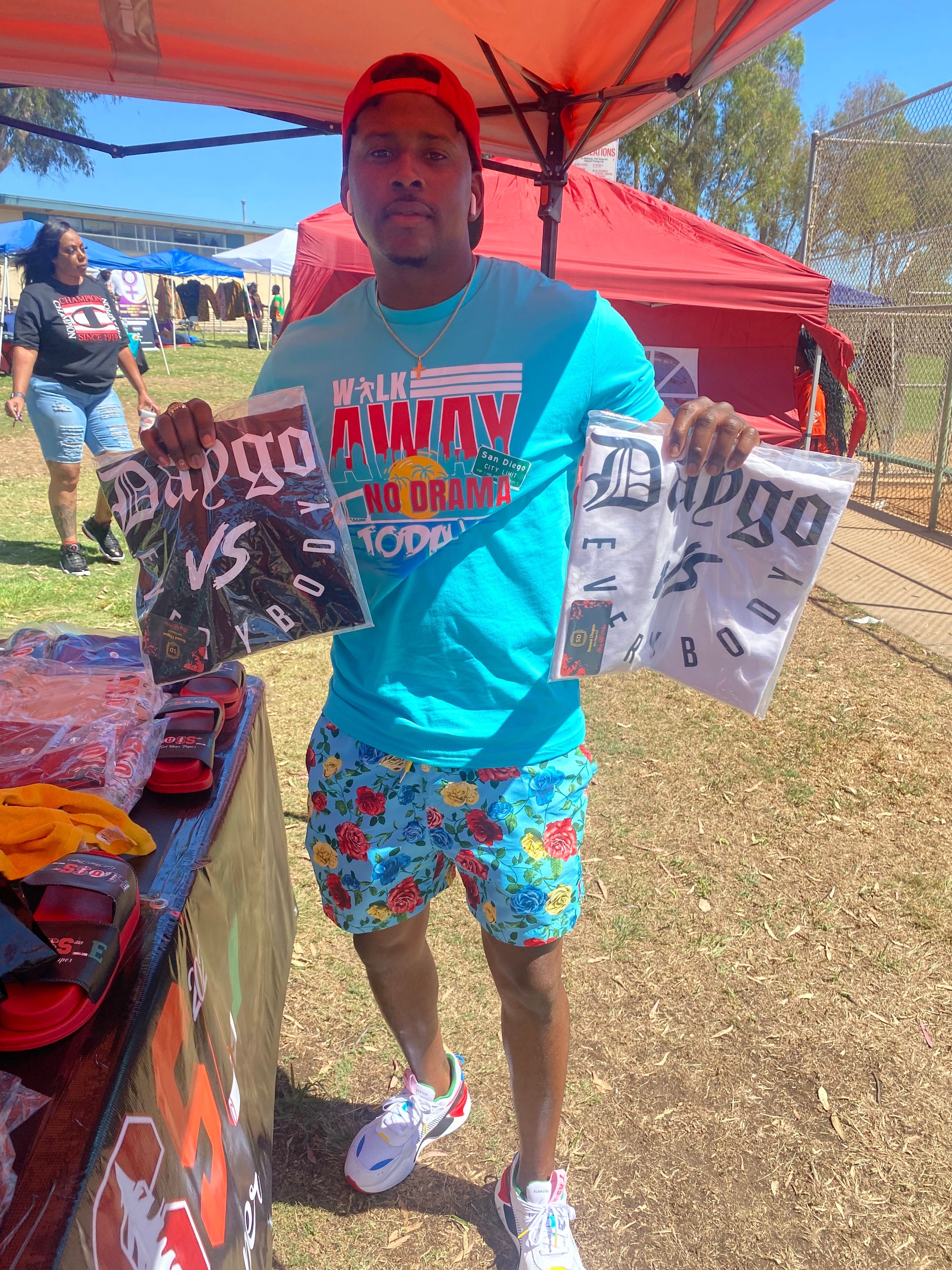 Man wearing a red cap, blue T-shirt, and colorful shorts holding two shopping bags outdoors.
