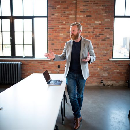 Man with beard in blazer and jeans standing in a bright office space with large windows and brick wall, gesturing.