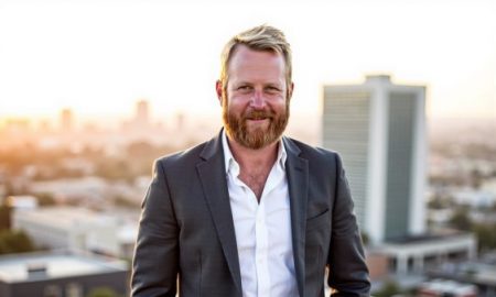 Man with a beard in a suit standing on a rooftop with cityscape background during sunset.