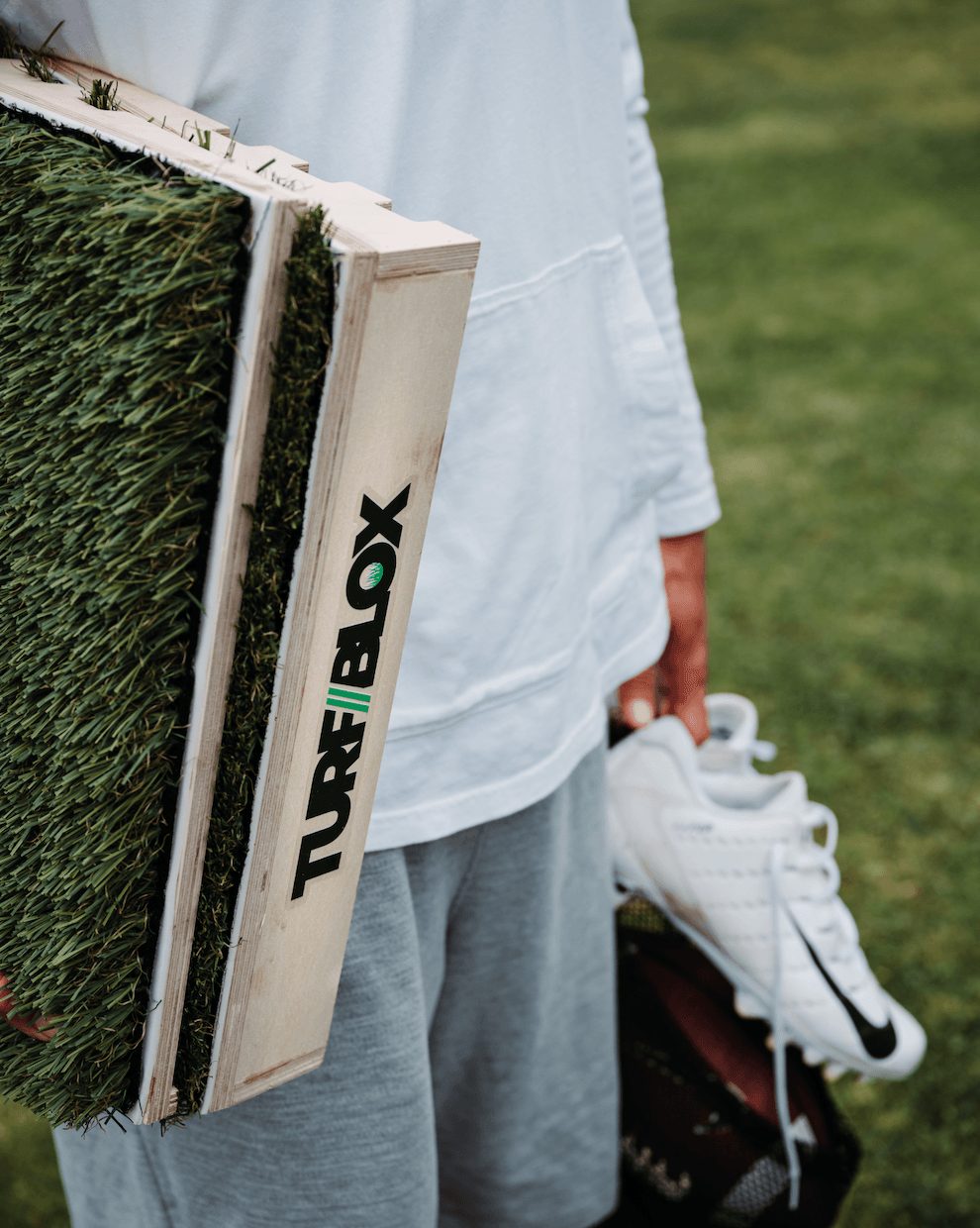Person holding a shoe and a wooden box with artificial grass outdoors, wearing a light hoodie and gray pants.