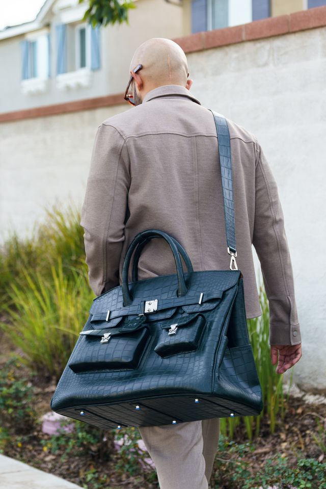 Bald person with glasses carrying a large blue bag, walking outdoors near a building and greenery.