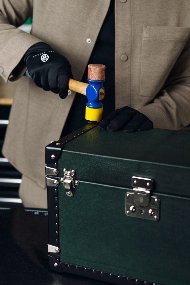 Person in suit and gloves using a hammer on a green box with metal latches.