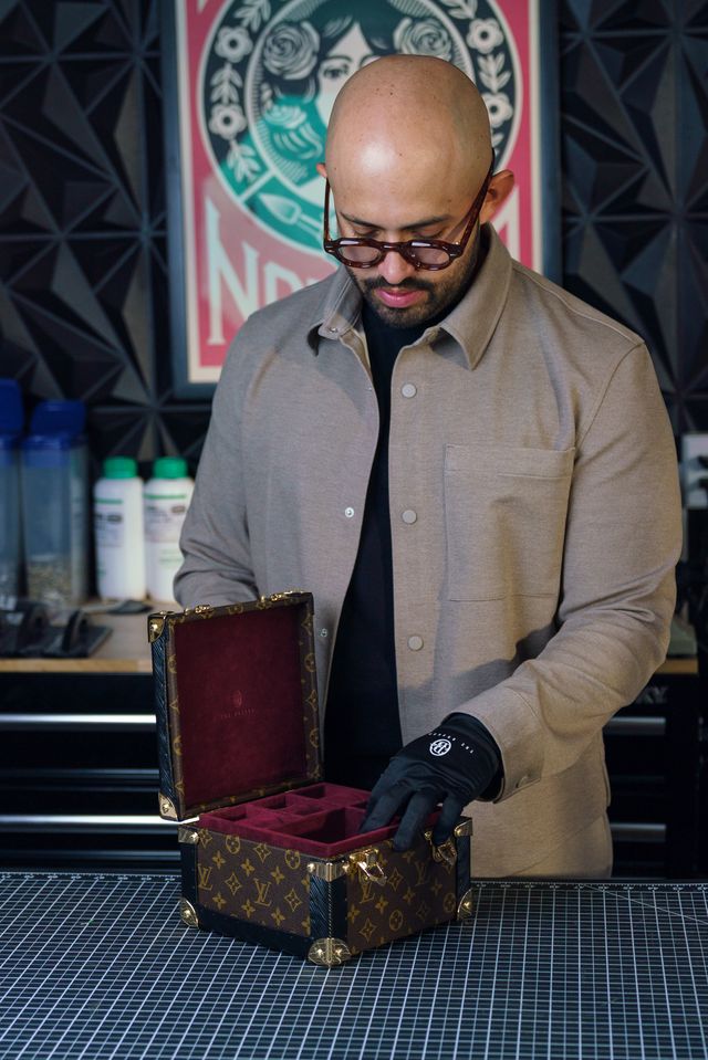 Bald man with glasses opening a decorated box on a table, wearing a beige coat and black gloves.