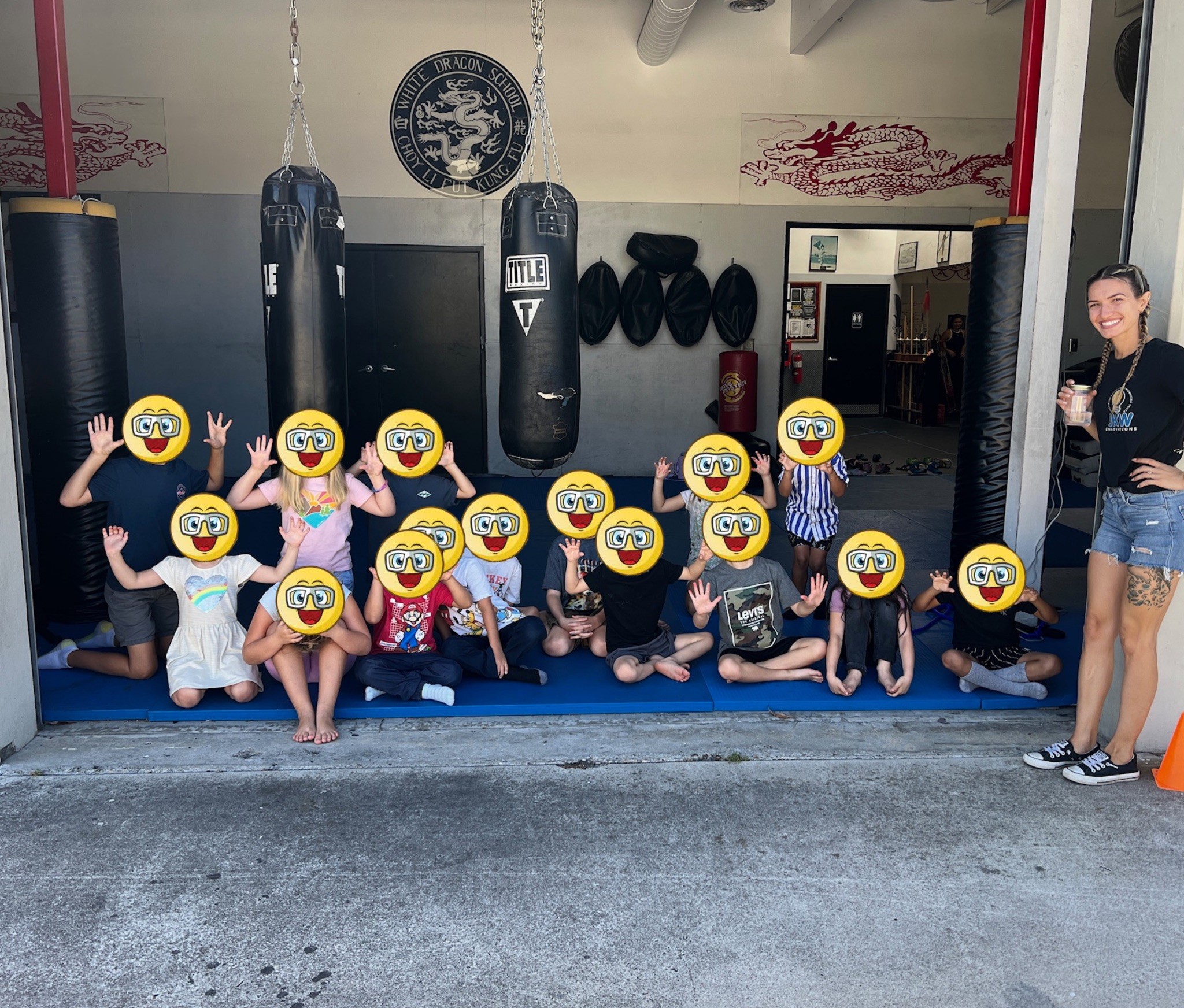 Group of children with emoji faces sitting on the floor in a gym, with a woman standing to the right.