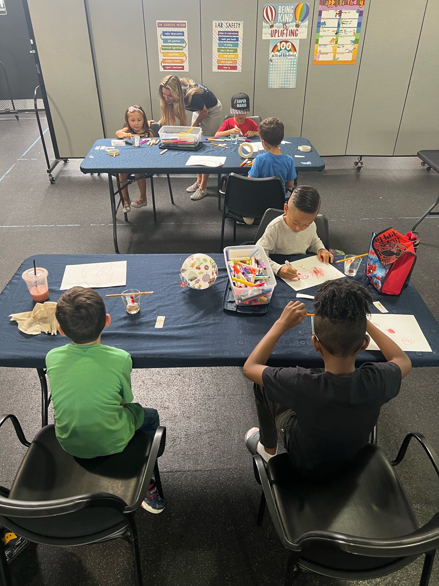 Children sitting at tables in a classroom, engaging in arts and crafts activities, with colorful posters on the wall behind.
