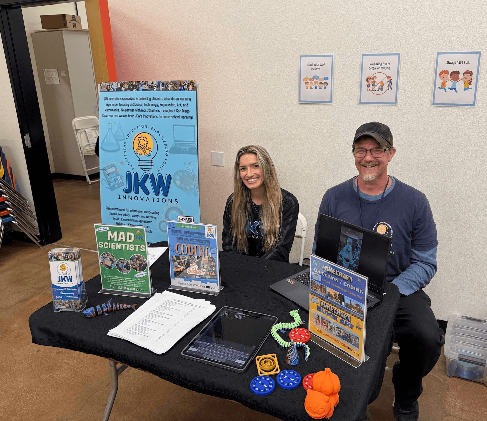 Two people sitting at a table with educational materials and posters, smiling in an indoor setting.