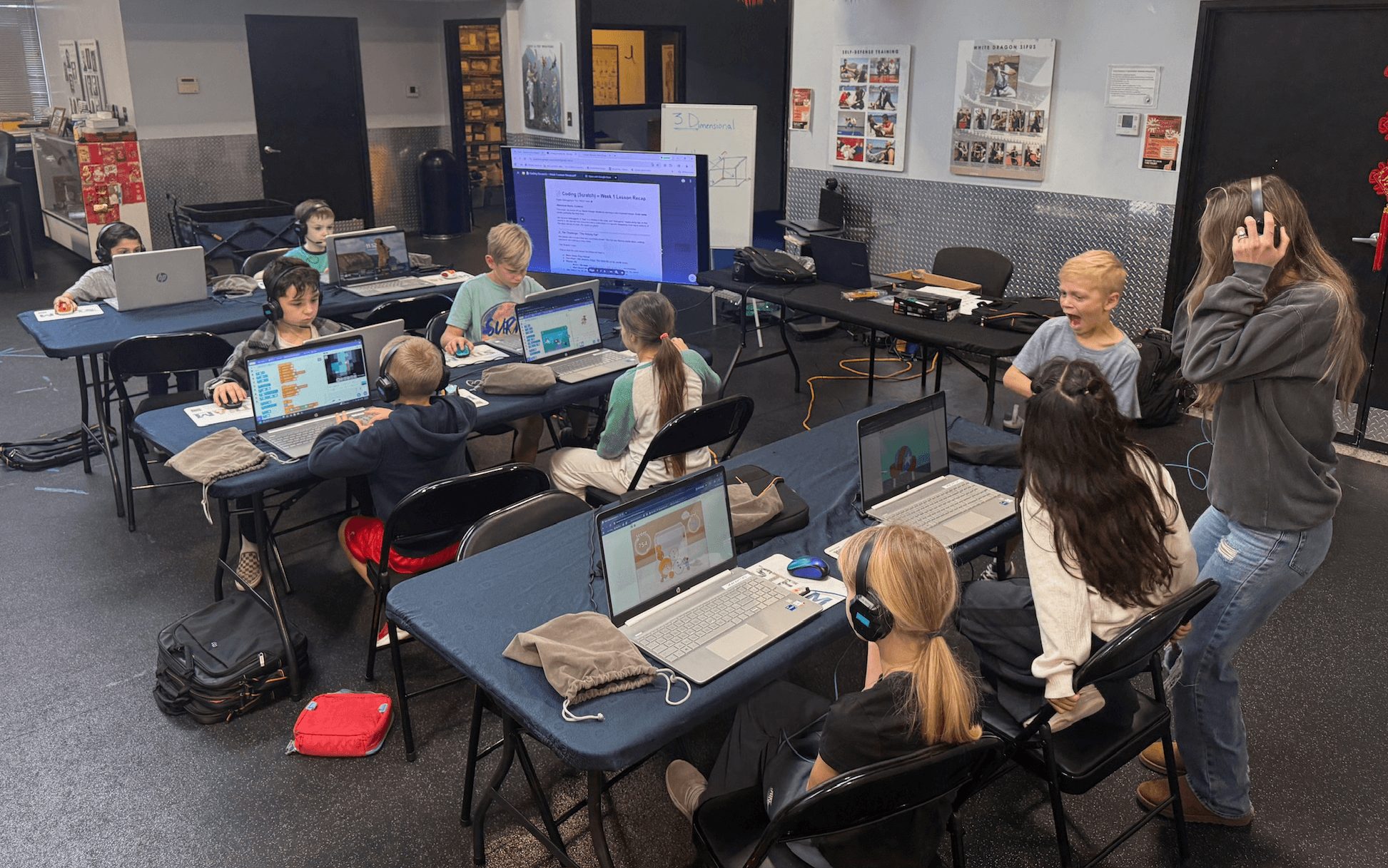 Classroom with students working on laptops at desks, teacher standing near whiteboard, and a large screen at the front.