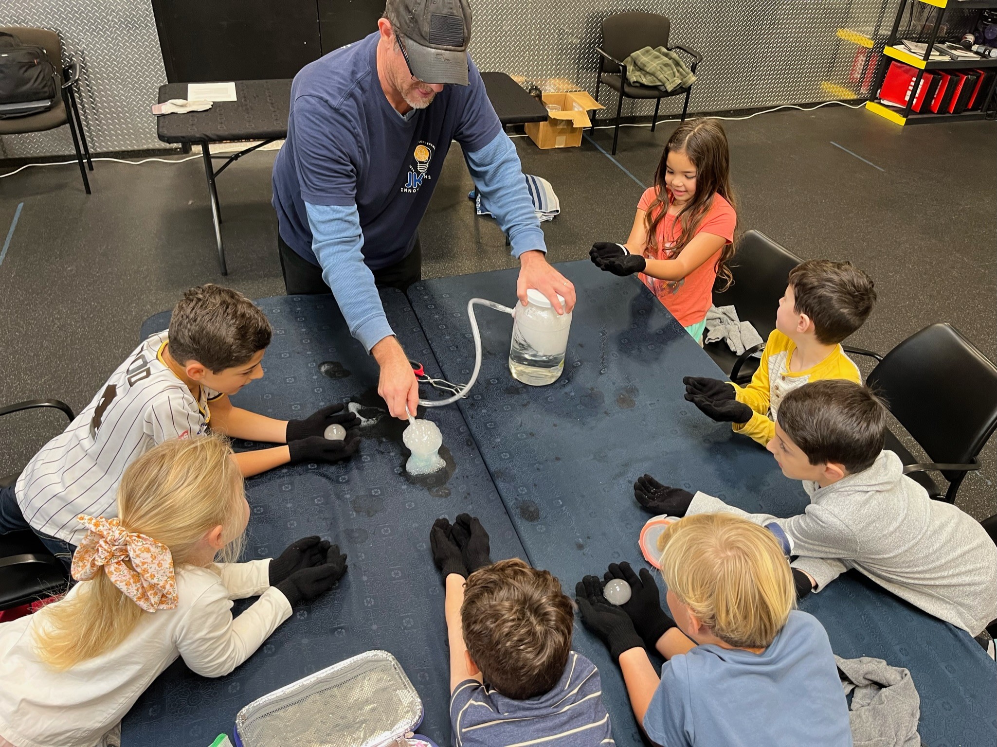 Adult demonstrating science experiment to children seated around a table in a classroom.