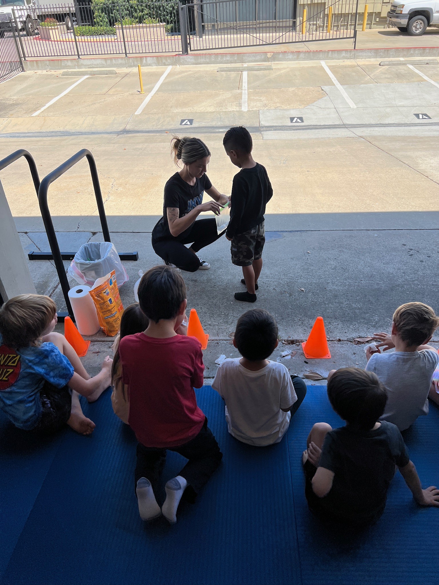Woman kneeling and talking to a child outside, with children sitting on the ground nearby and orange cones in front.