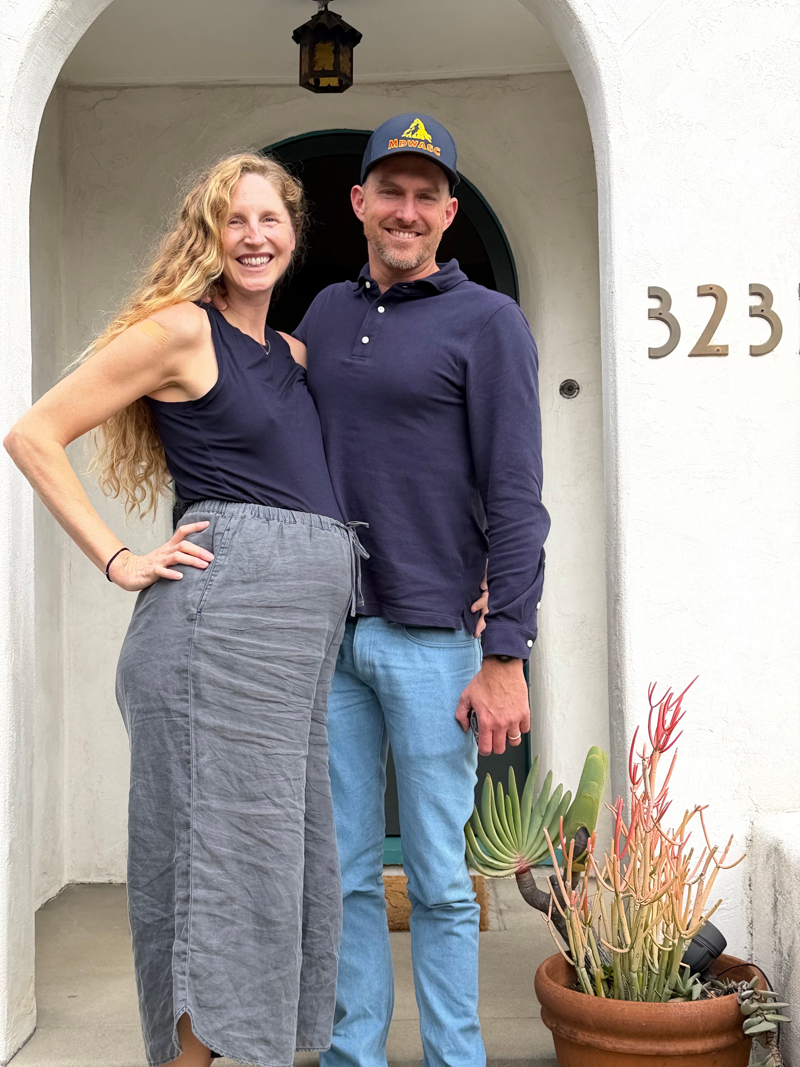 A smiling woman with long curly hair and a man with a cap stand together outside a white building with the number 323, plants nearby.