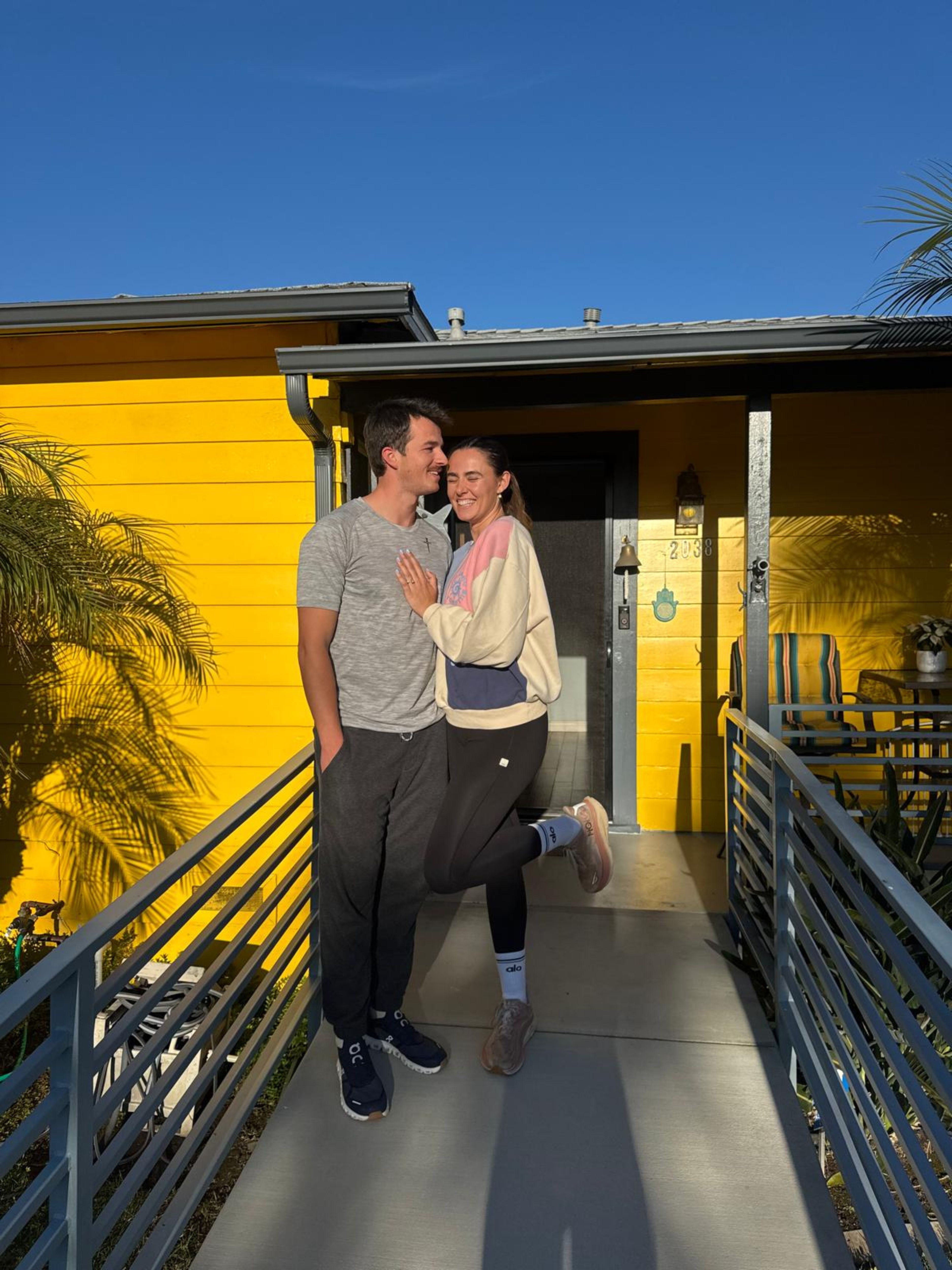Two people standing on a walkway in front of a yellow building, smiling and posing together.