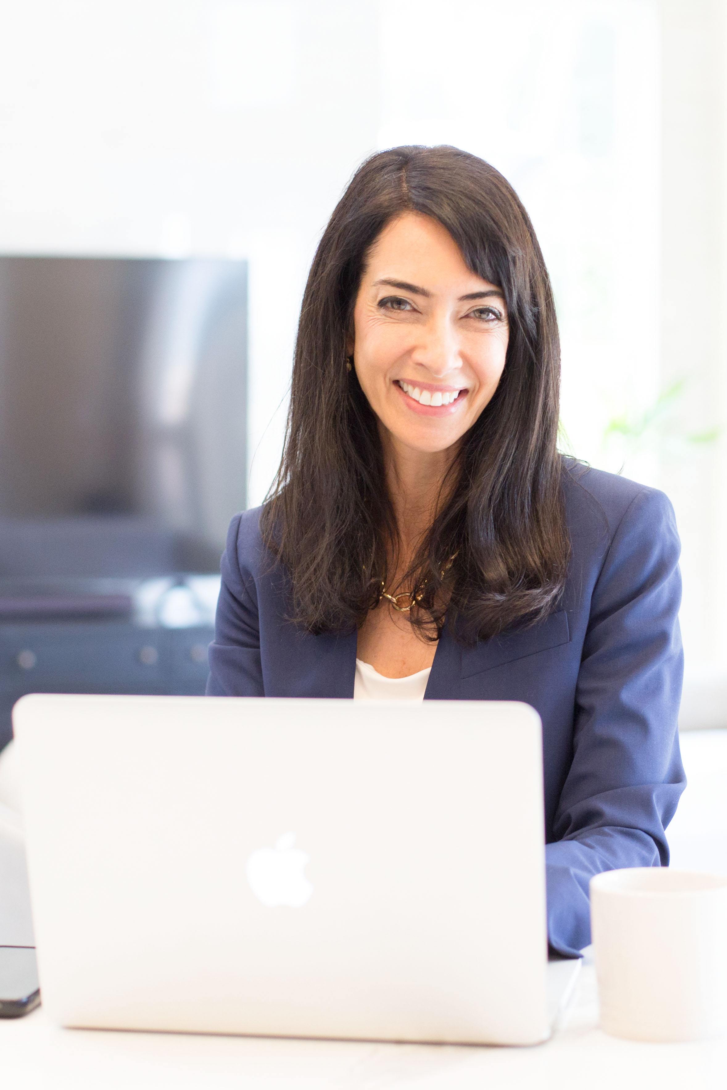 Woman with dark hair smiling, sitting at desk with laptop, in office environment.