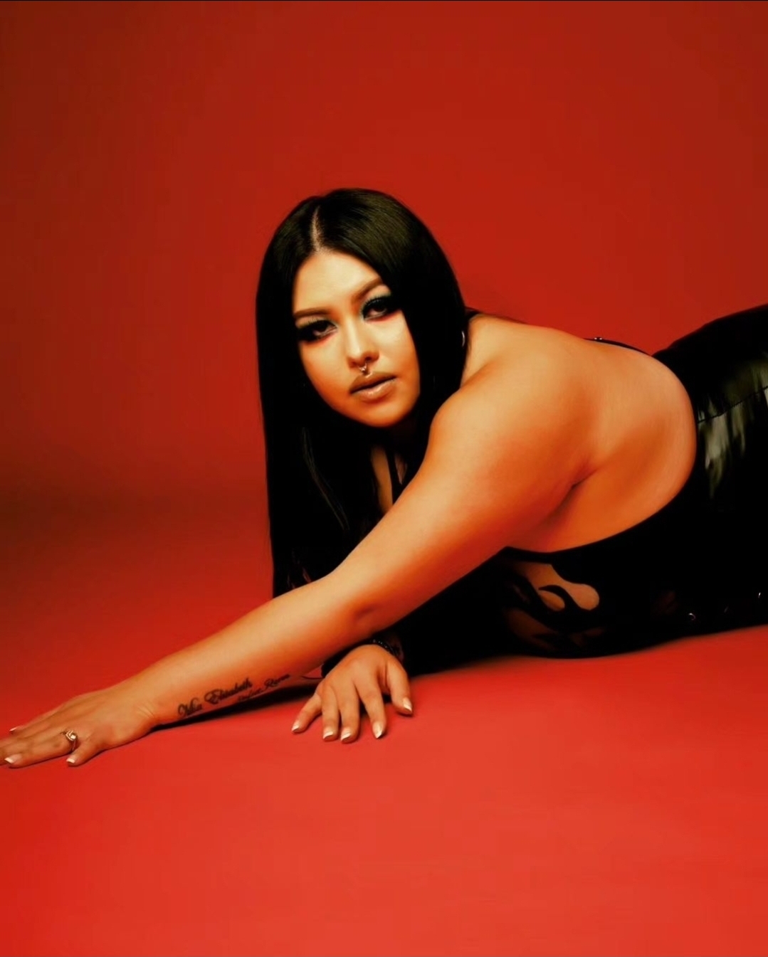 Woman with long black hair lying on red surface, looking at camera, wearing black outfit, against red background.