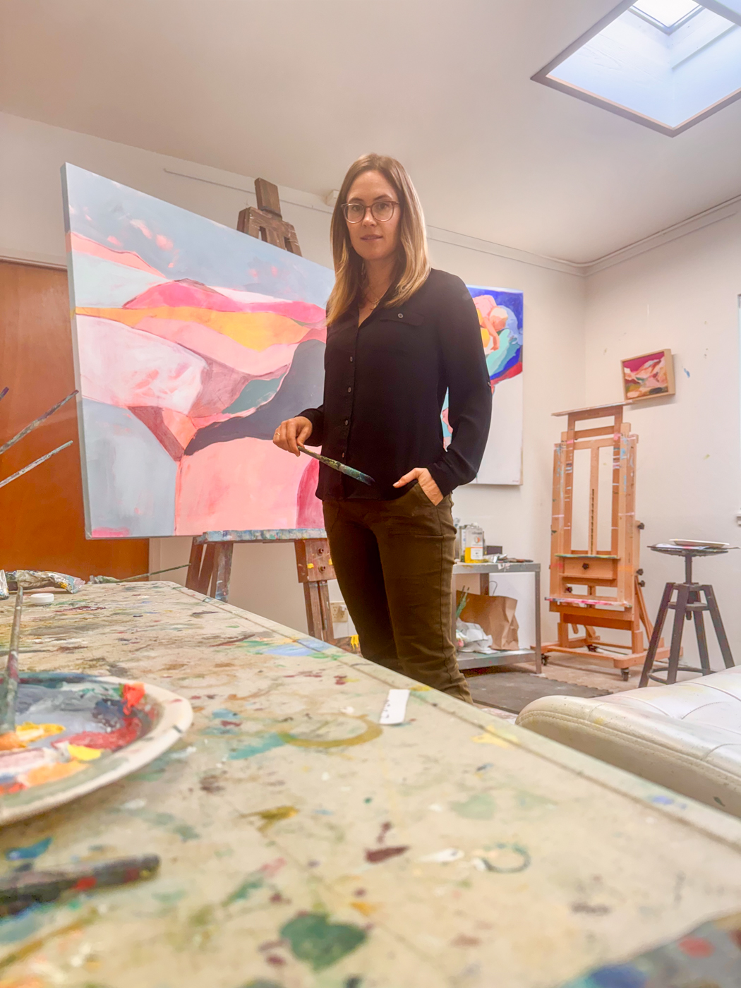 Woman standing in art studio with paintings and art supplies, holding a palette, wearing glasses and dark clothing.