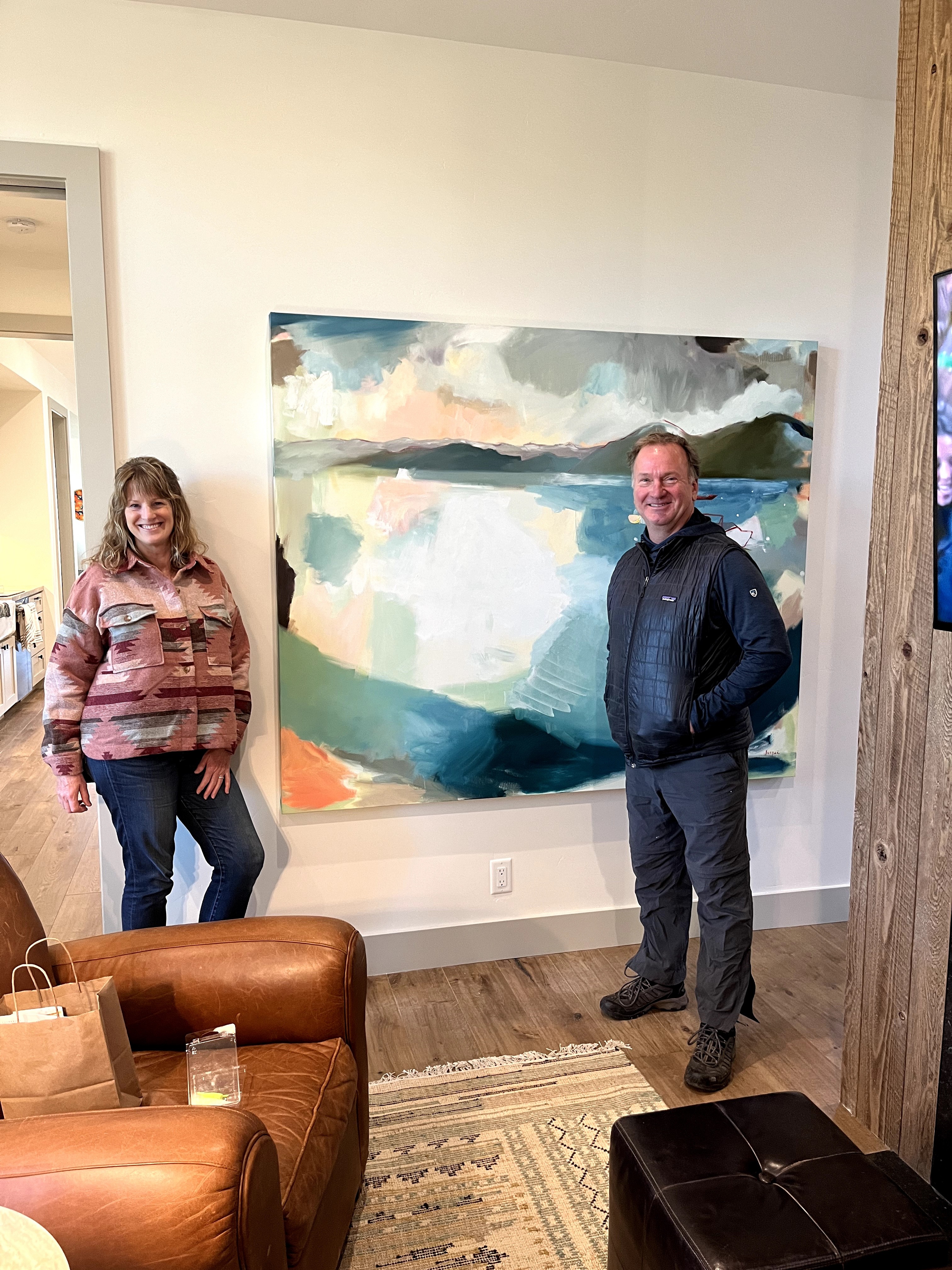 Two people stand in front of a large abstract landscape painting on a wall, with a leather chair and wooden floor nearby.