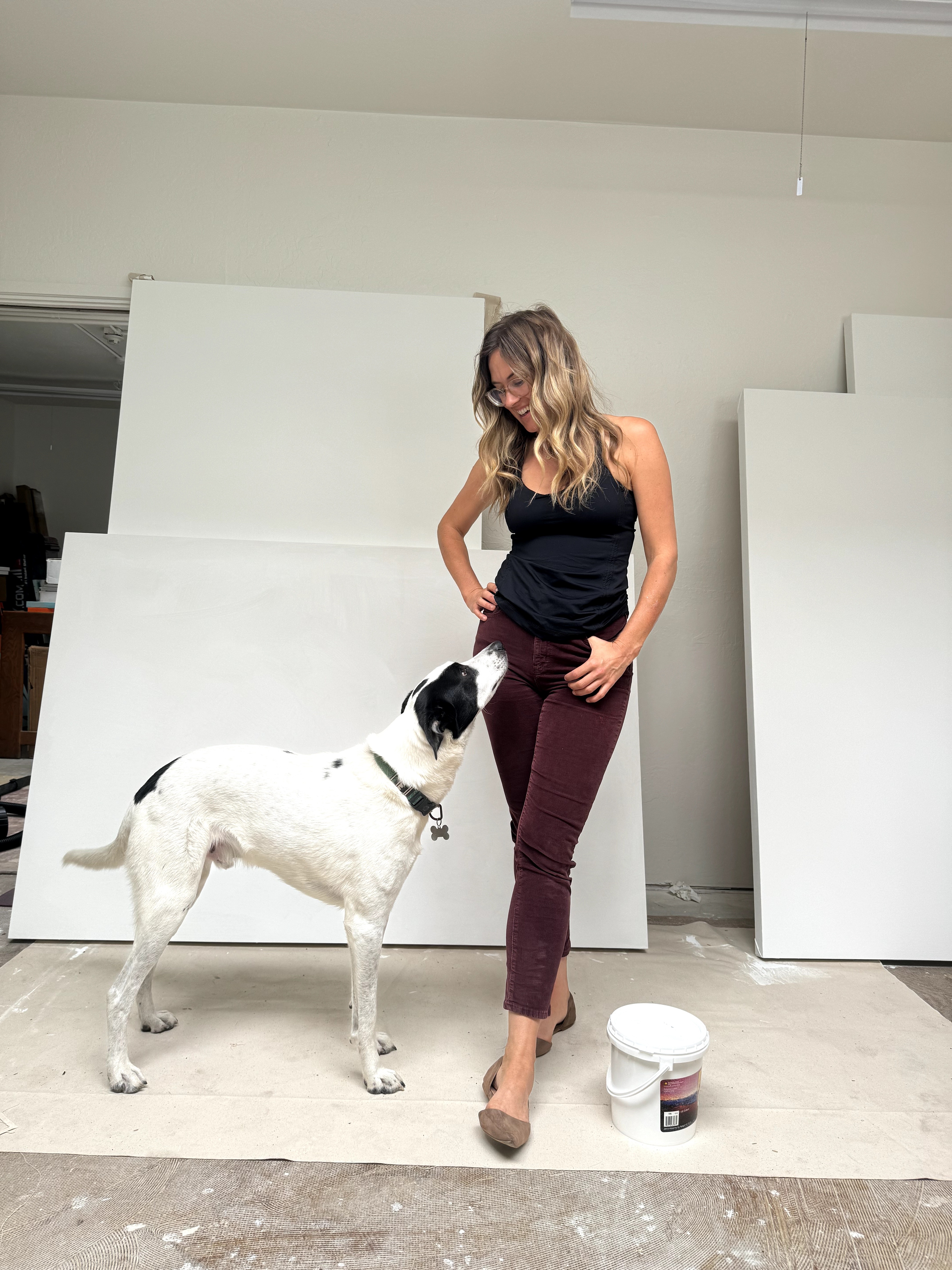 Woman with wavy hair in black top and maroon pants standing next to a large white and black dog indoors.