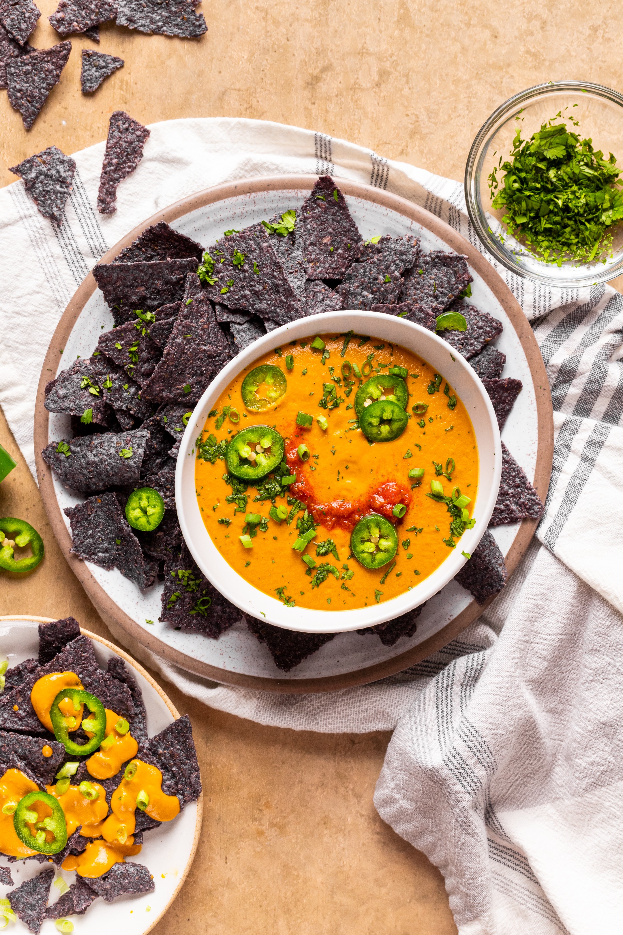 Bowl of orange-colored soup with green jalapeños and herbs, surrounded by black chips and a small bowl of chopped herbs.