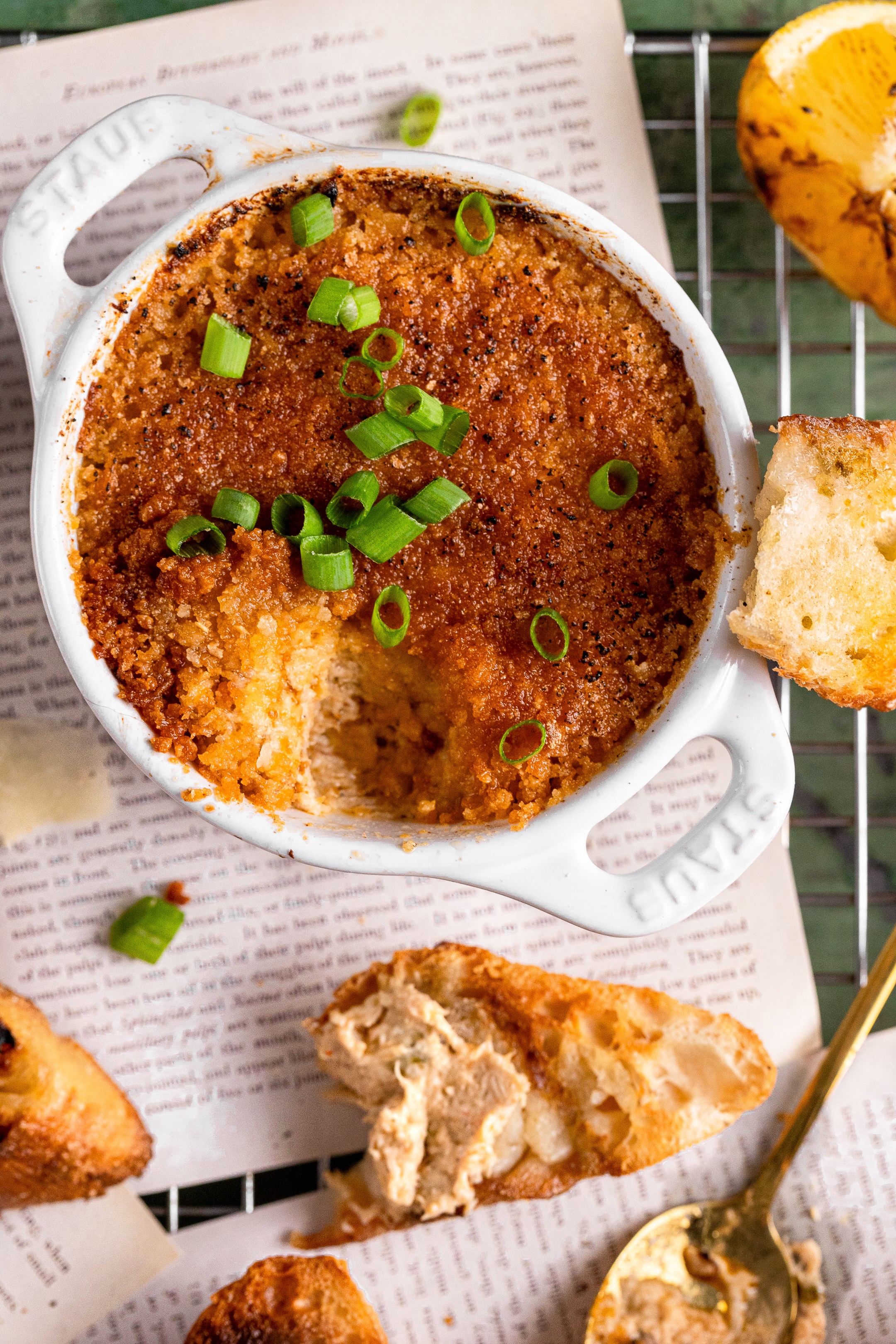 Baked dish with green onions in a white bowl, bread on side, spoon nearby, on newspaper-lined surface.
