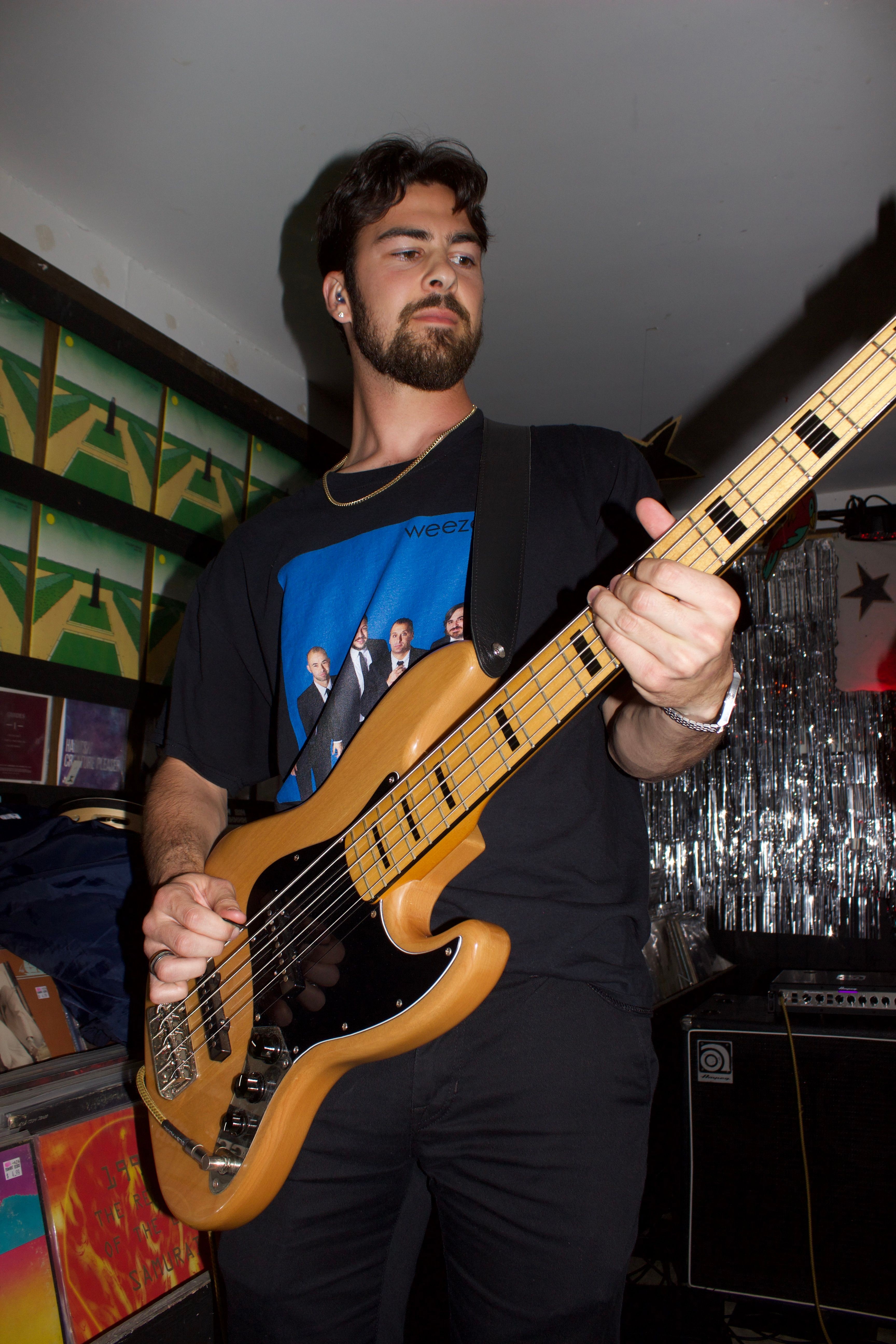 Young man with dark hair and beard playing a bass guitar indoors, wearing a black T-shirt with a graphic design.