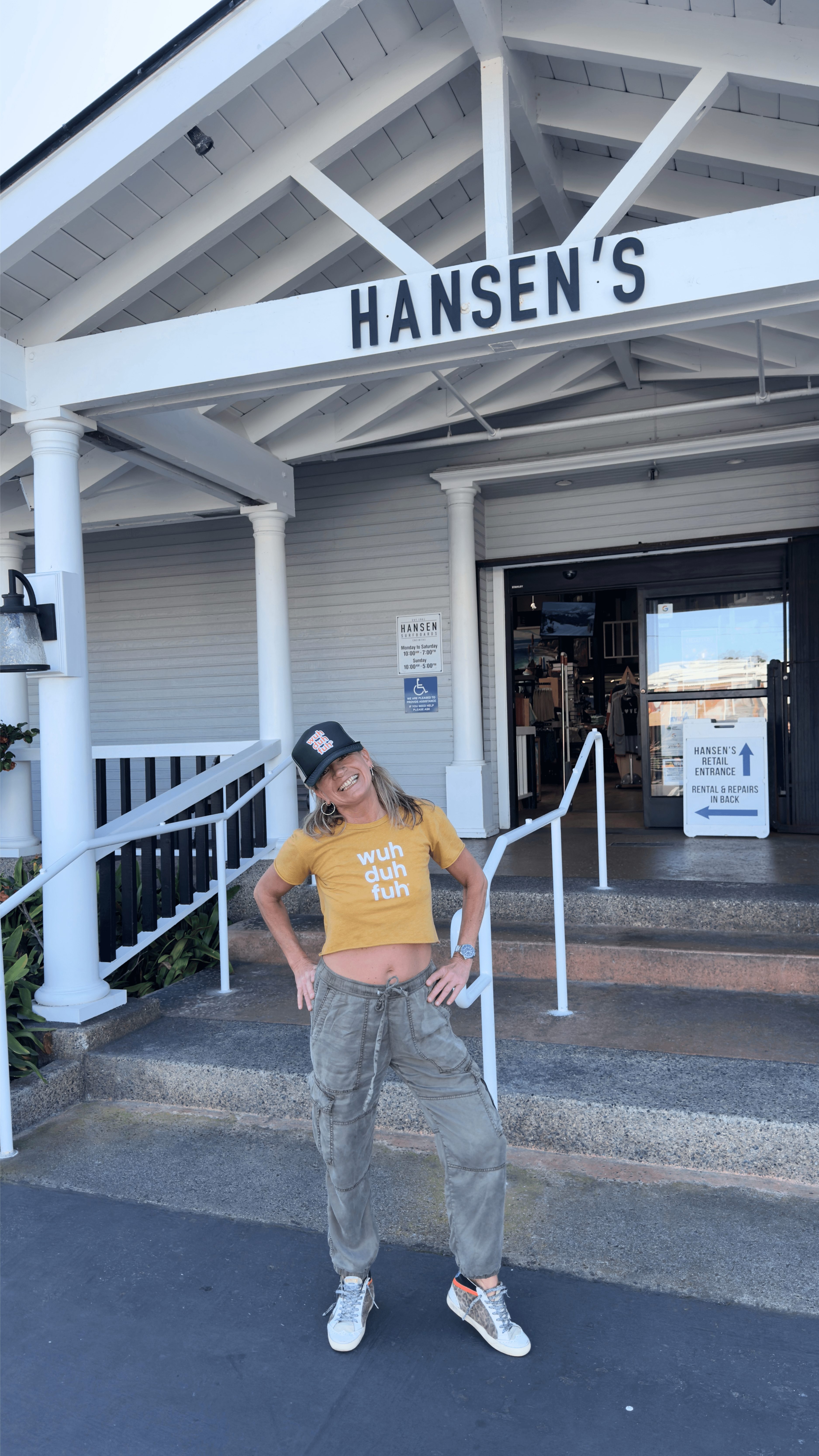 Person standing outside Hansen's store, wearing a yellow shirt, gray pants, and a black cap, smiling at camera.