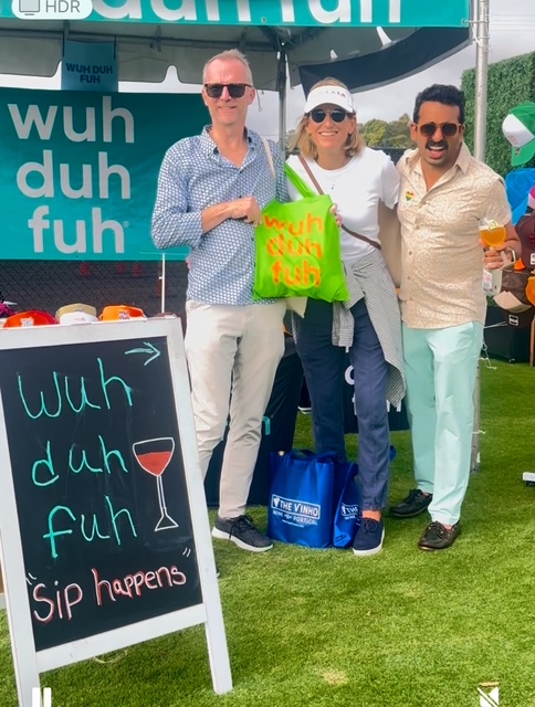 Three people standing outdoors near a sign and a chalkboard, holding drinks and a green bag, smiling.