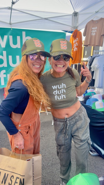 Two smiling women wearing hats and casual clothing, standing outdoors at a market stall with clothing and merchandise.