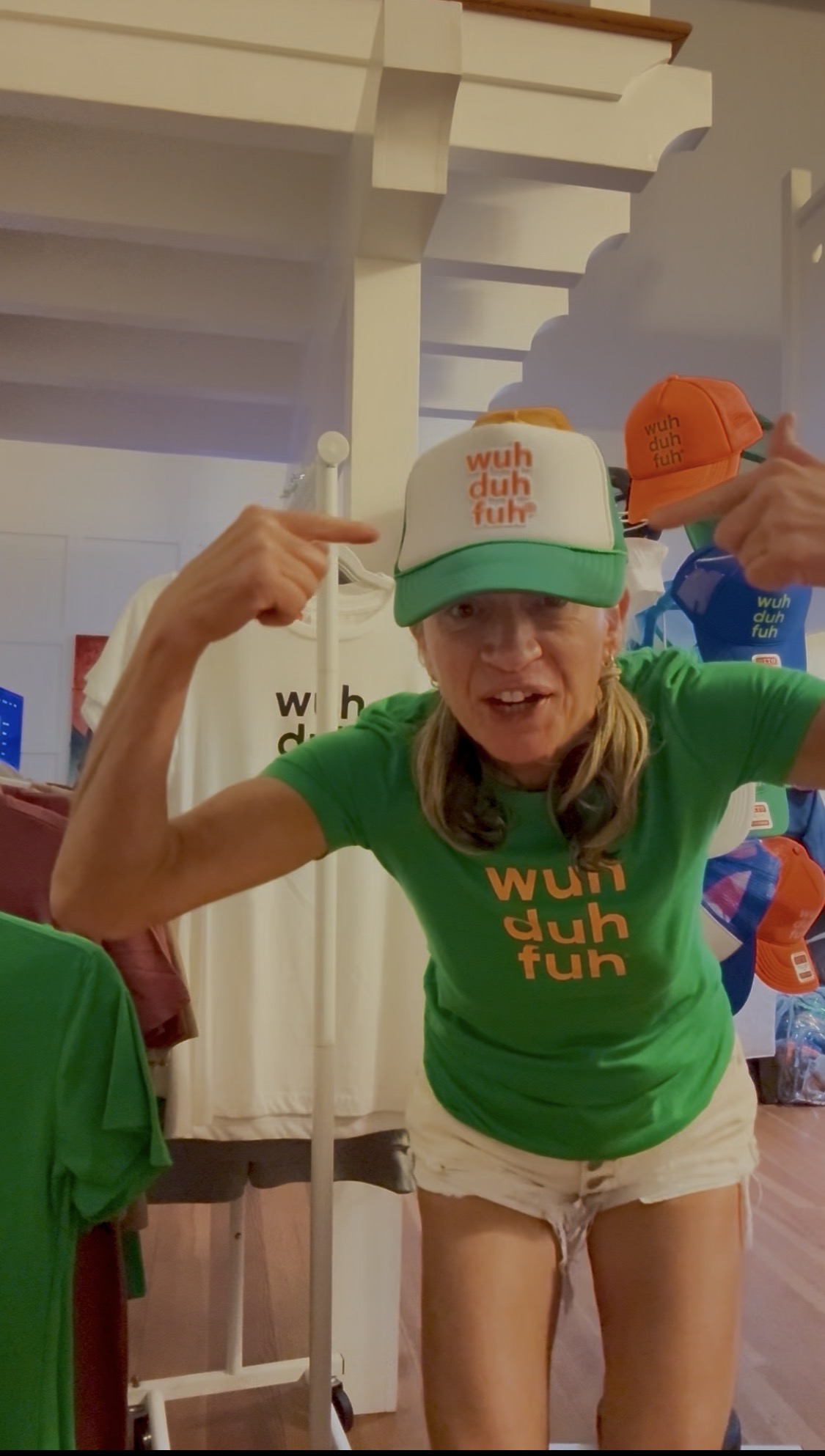 Woman wearing a green shirt and a trucker hat, pointing at her head, indoors with colorful items around.