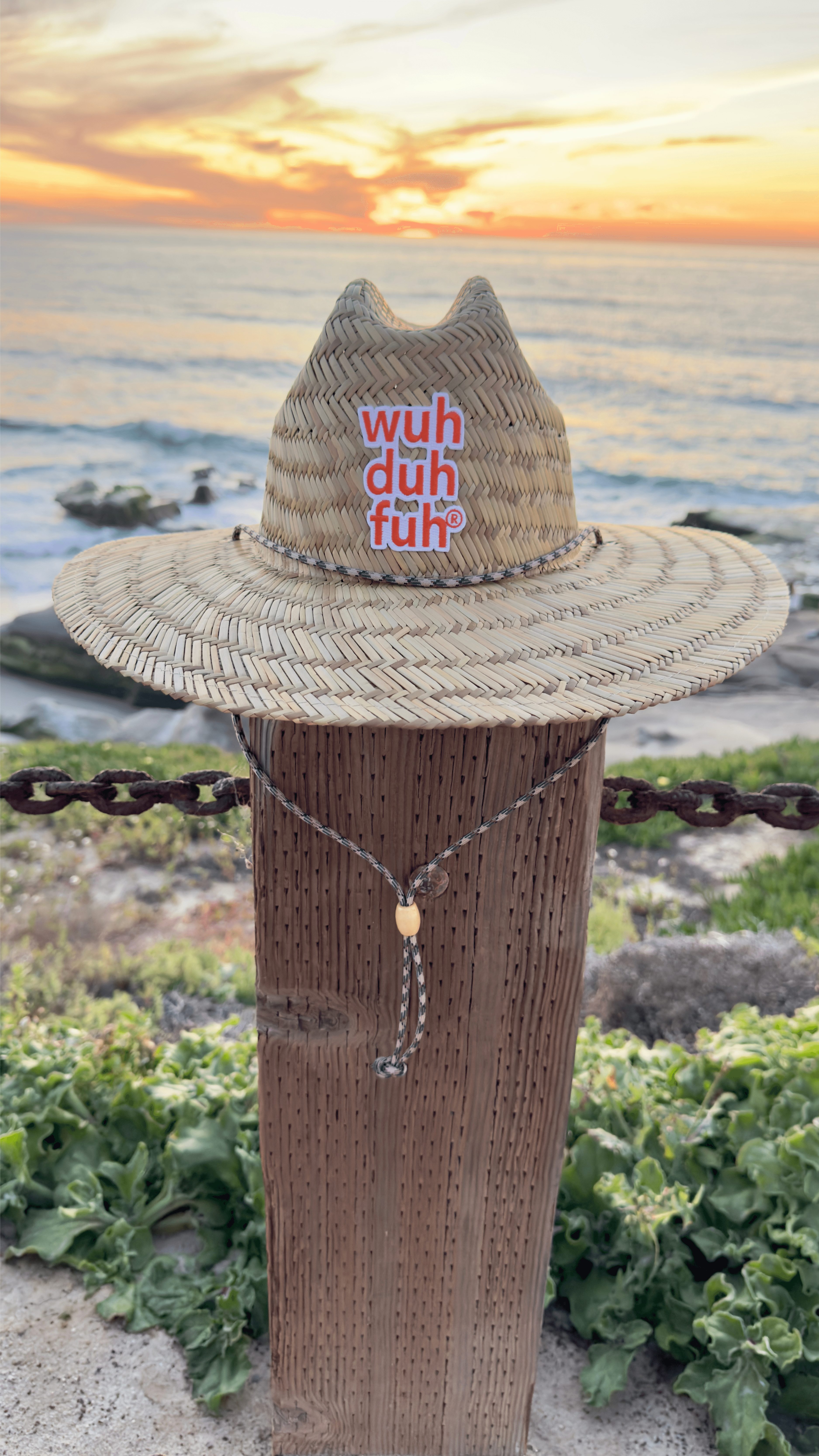 Straw hat with pink text, placed on a wooden post near the ocean at sunset, with greenery in foreground.