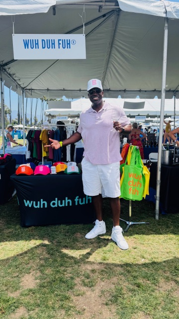 Man standing in front of a booth with colorful items, wearing a pink shirt, white shorts, and a white cap, outdoors.