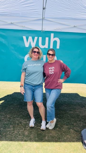 Two women stand outdoors in front of a blue banner with the word 'wuh'. They wear sunglasses and casual clothing.