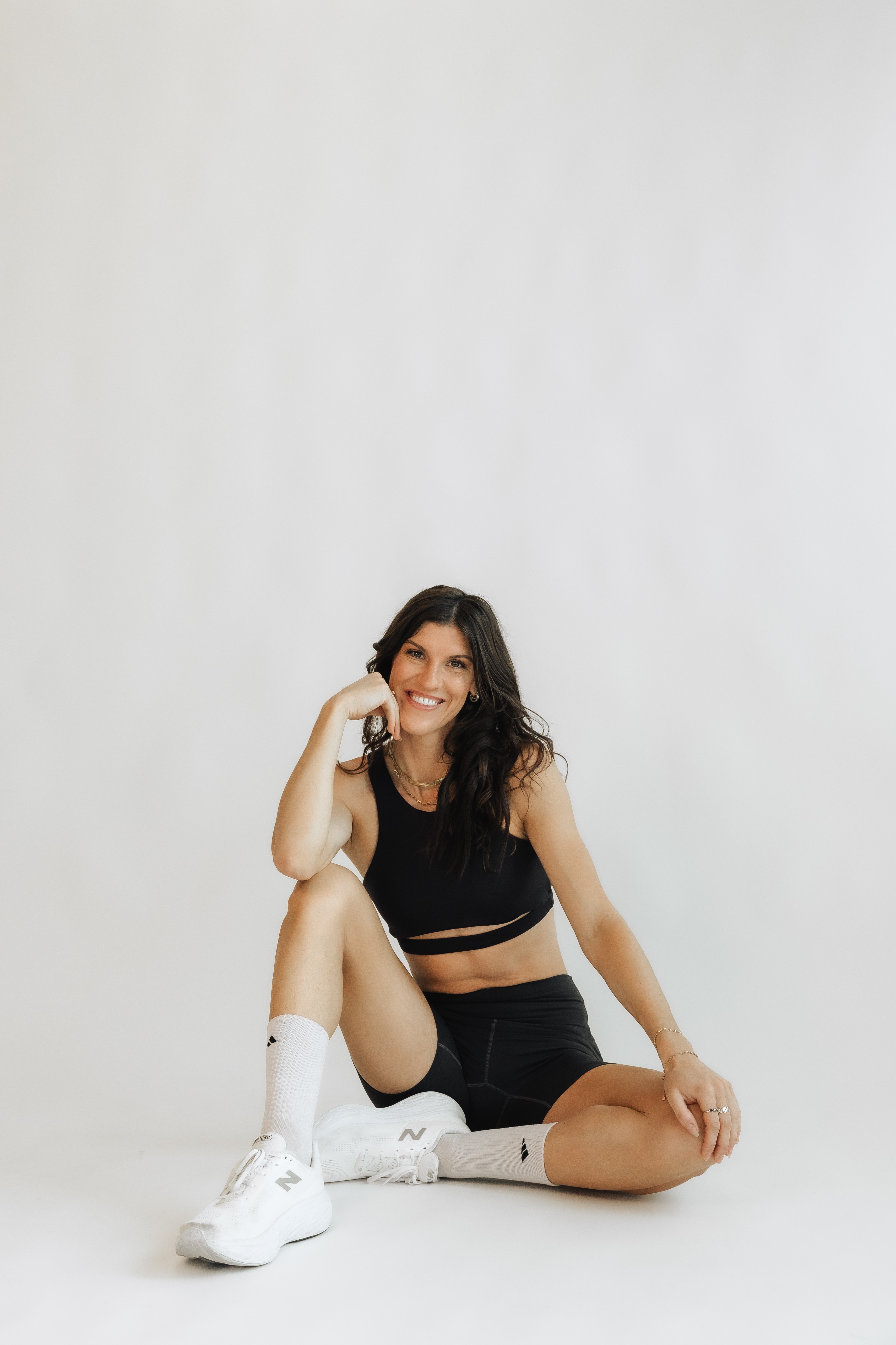 Young woman sitting on the floor, smiling, wearing black crop top, shorts, and white sneakers, against a plain white background.