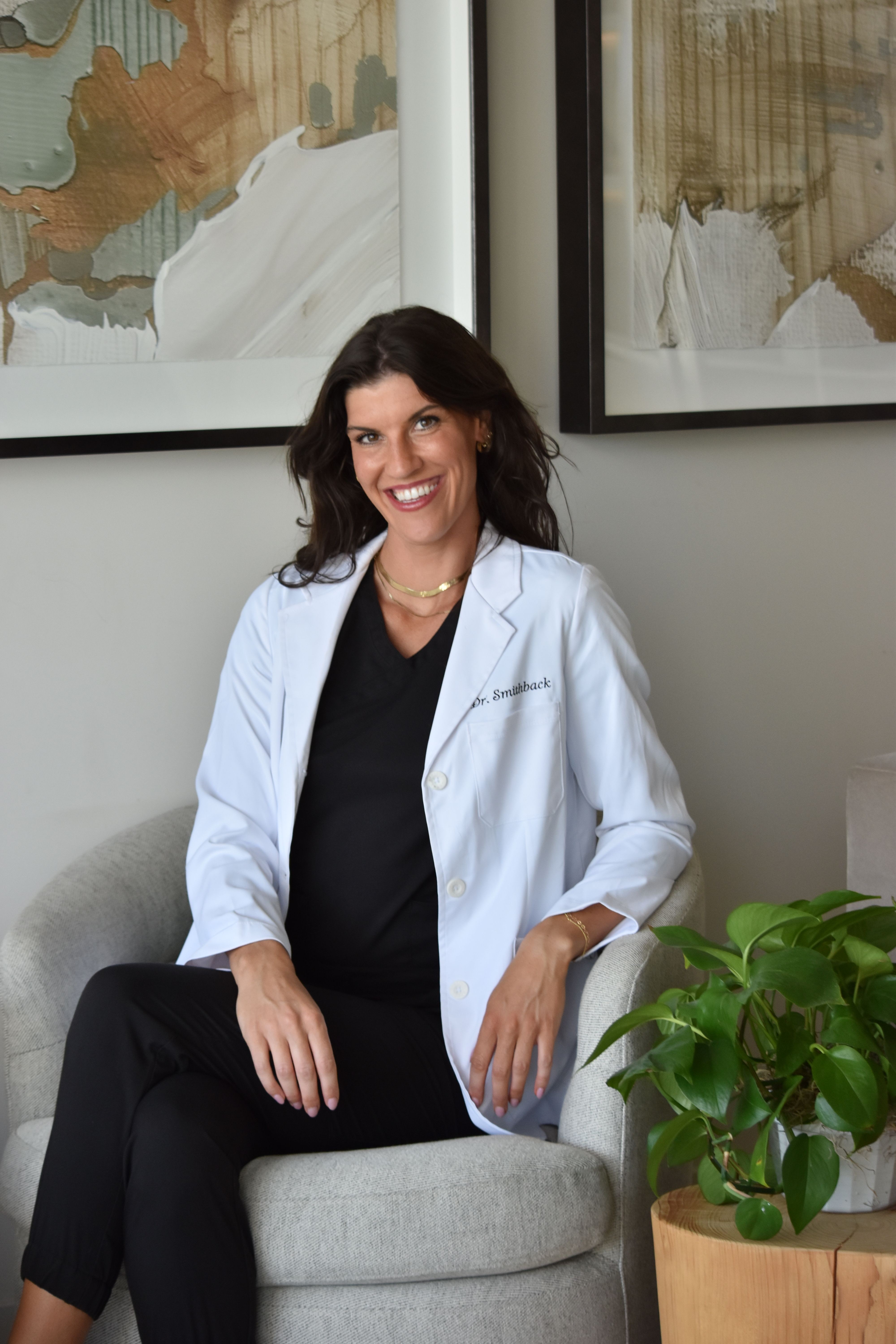 Woman in white coat sitting on gray chair, smiling, in a room with abstract artwork and a green plant.