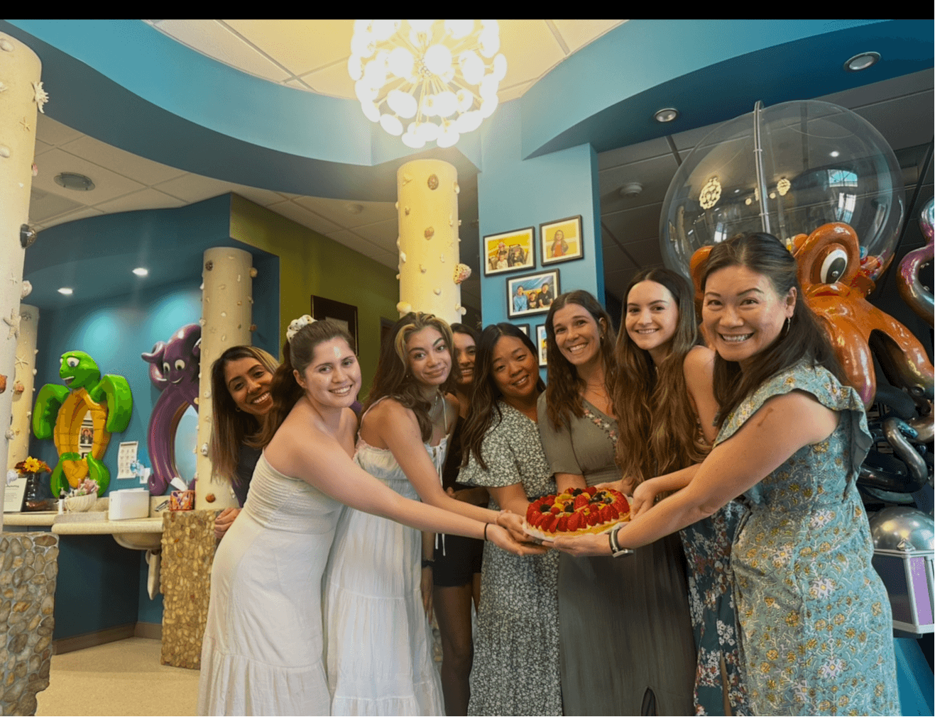Group of six women smiling and holding a cake in an indoor setting with colorful decorations.