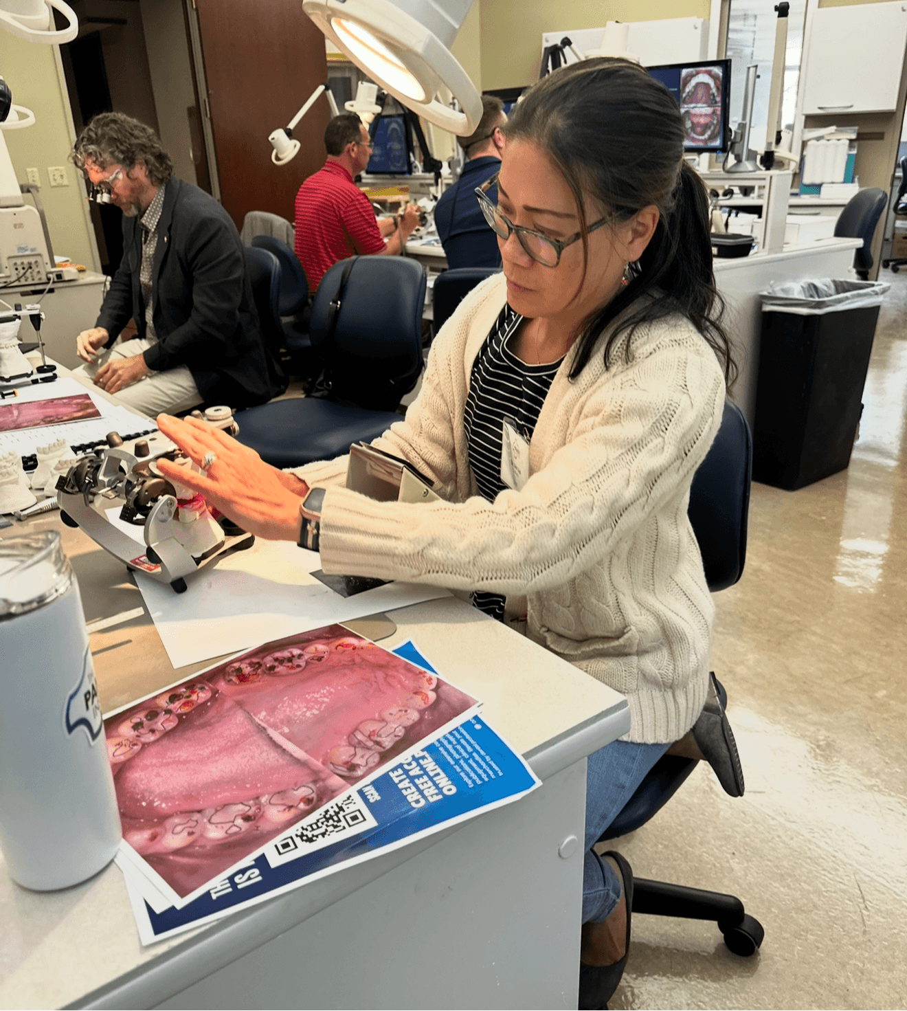 Woman in glasses and beige sweater working with medical equipment at a desk in a laboratory.
