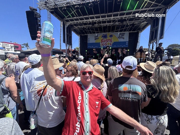 Crowd gathered at an outdoor concert with a stage and a woman holding a water bottle, smiling, in the foreground.