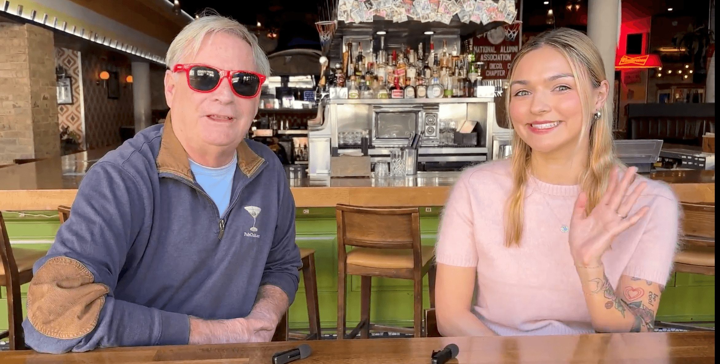 Two people sitting at a bar, smiling, with a bar counter and bottles in the background.