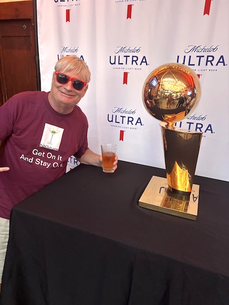 Person with sunglasses and a maroon shirt sitting at a table with a trophy and a drink, backdrop with 'Michelob ULTRA' logo.