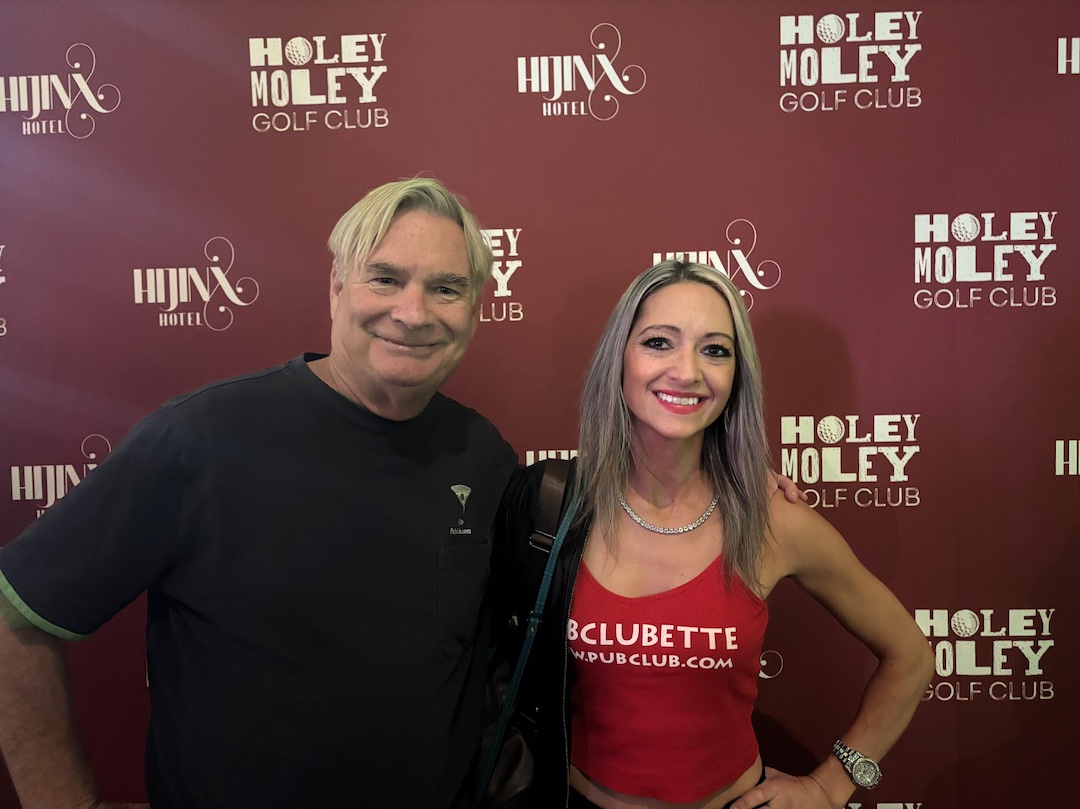Two people smiling in front of a red backdrop with logos for Holey Moley Golf Club and Hux Hotel.