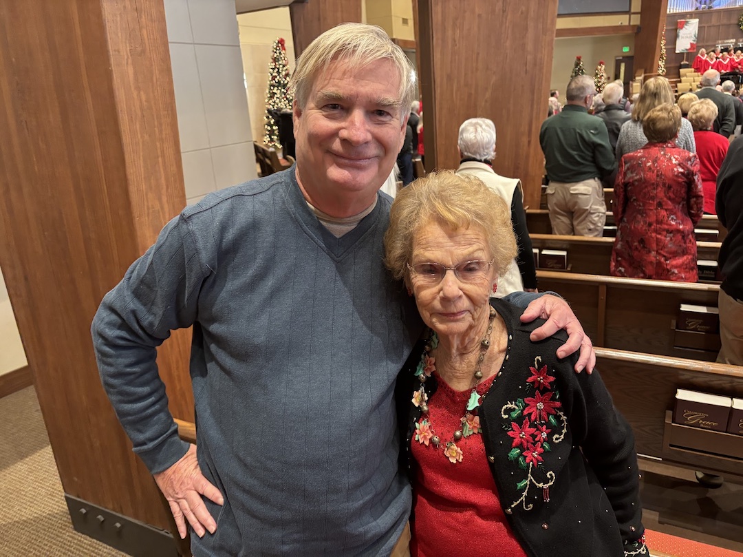 A man and an elderly woman pose together inside a church, with people seated in the background.