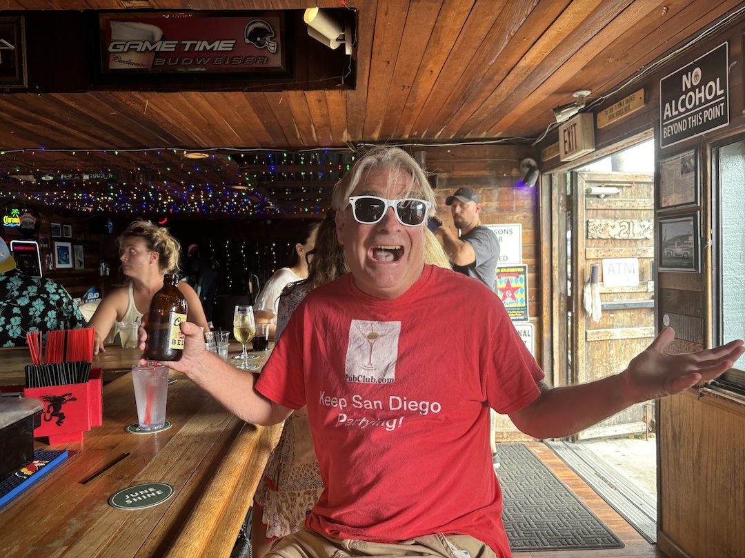 Person wearing sunglasses and a red shirt with arms outstretched inside a bar, smiling.