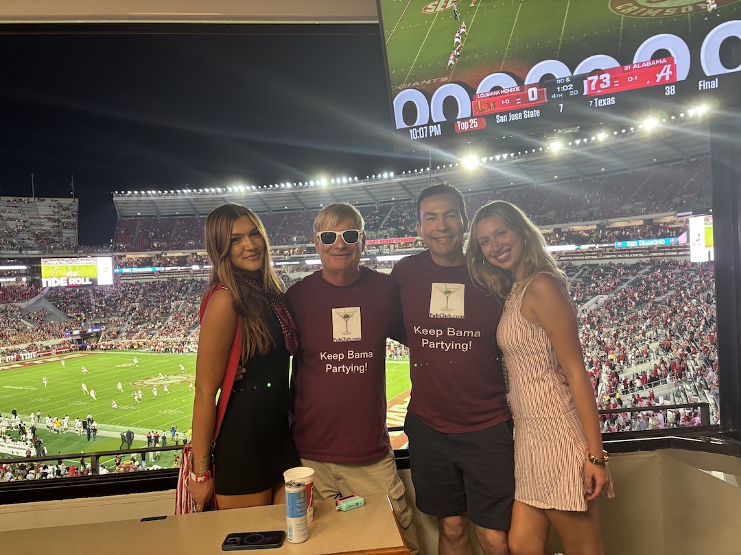 Four people standing together in a stadium, with a football game in progress behind them, smiling at the camera.