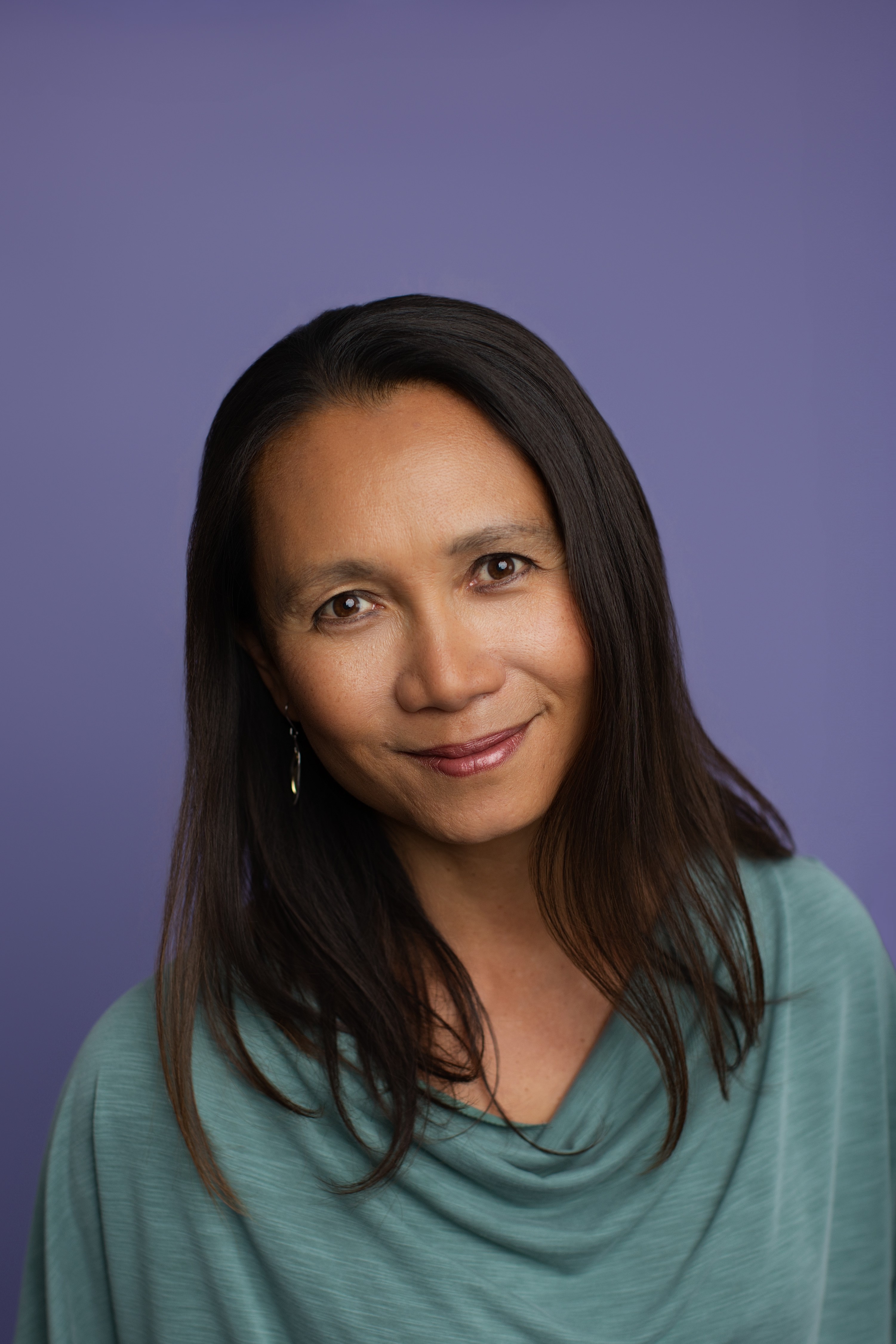 Woman with long dark hair smiling, wearing a teal top, against a purple background.